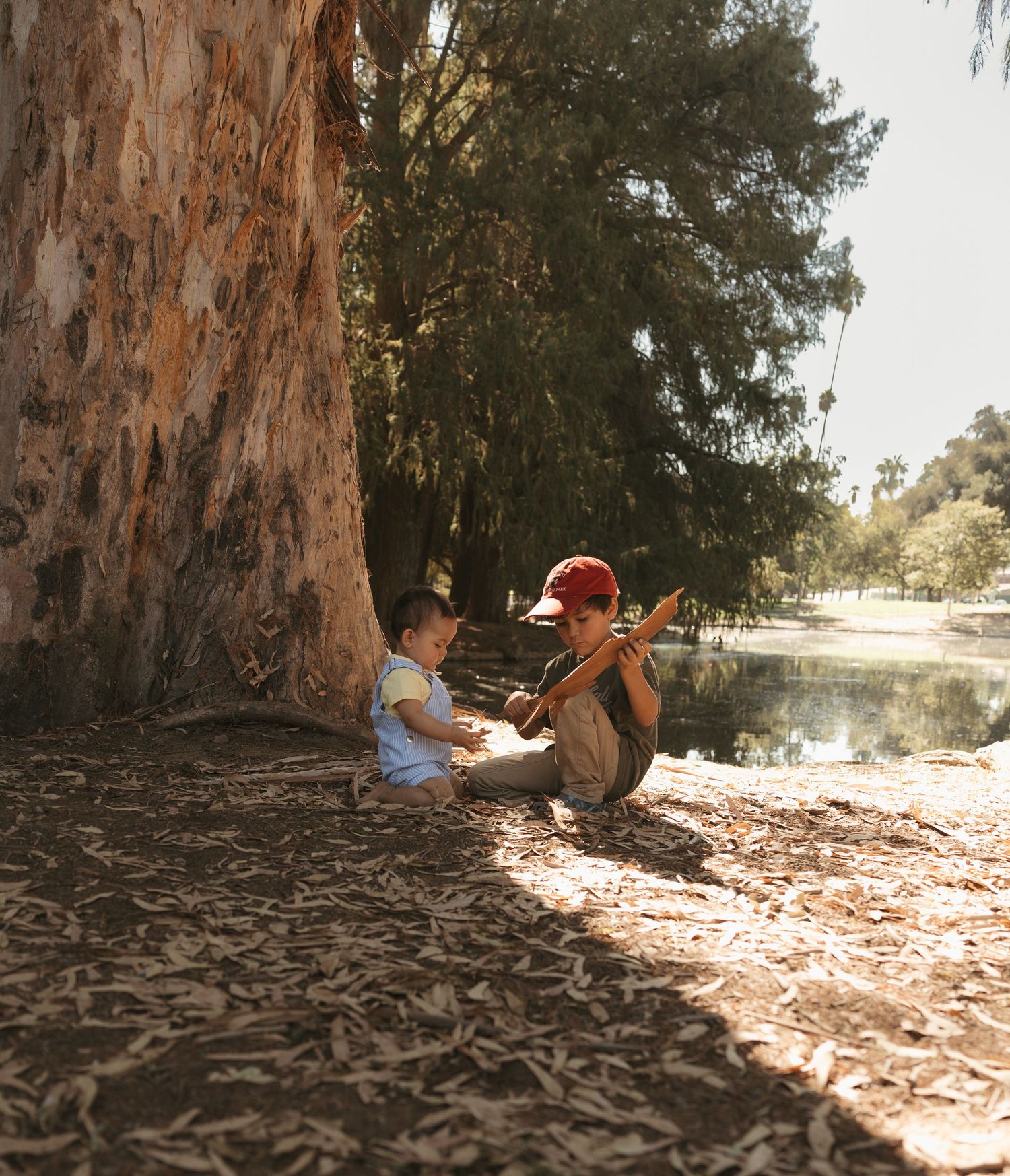 Two children sitting under a large tree near a pond in a sunlit park, one wearing a red cap playing outdoors.