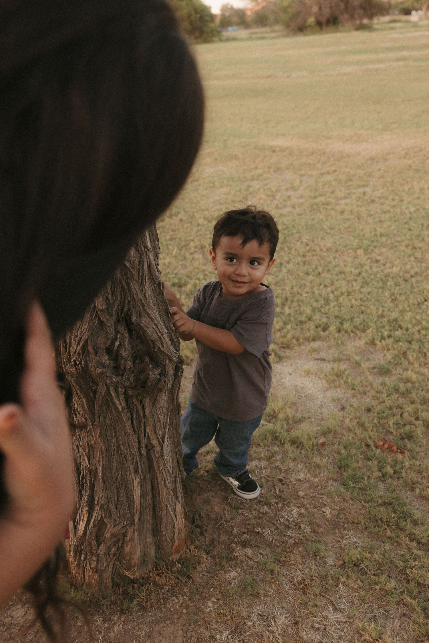 A young toddler boy stands beside a tree trunk in a grassy park, smiling at the camera.