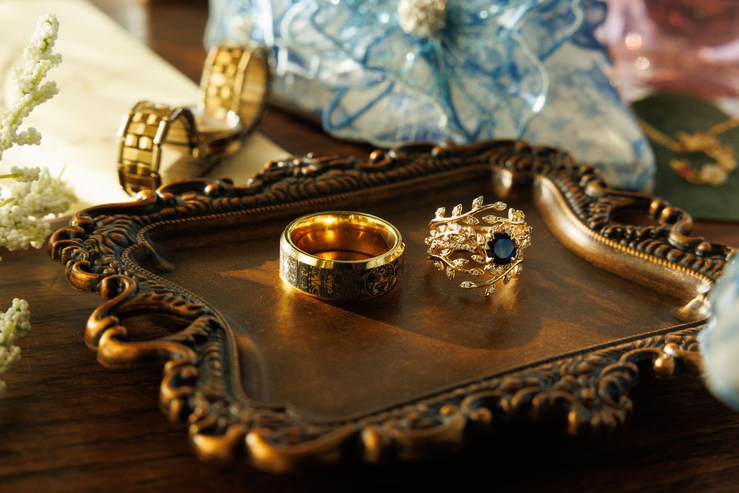 Ornate gold rings with blue gemstone displayed on antique decorative tray with floral accents.