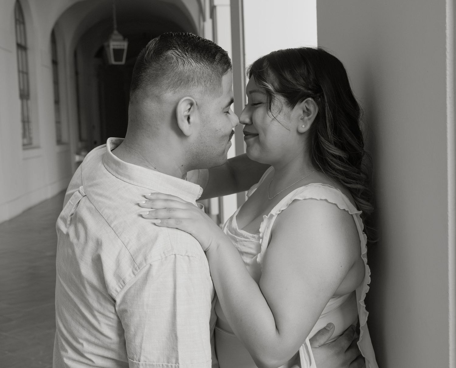 Black and white photo of a couple sharing a romantic moment, nearly kissing in a sunlit corridor.