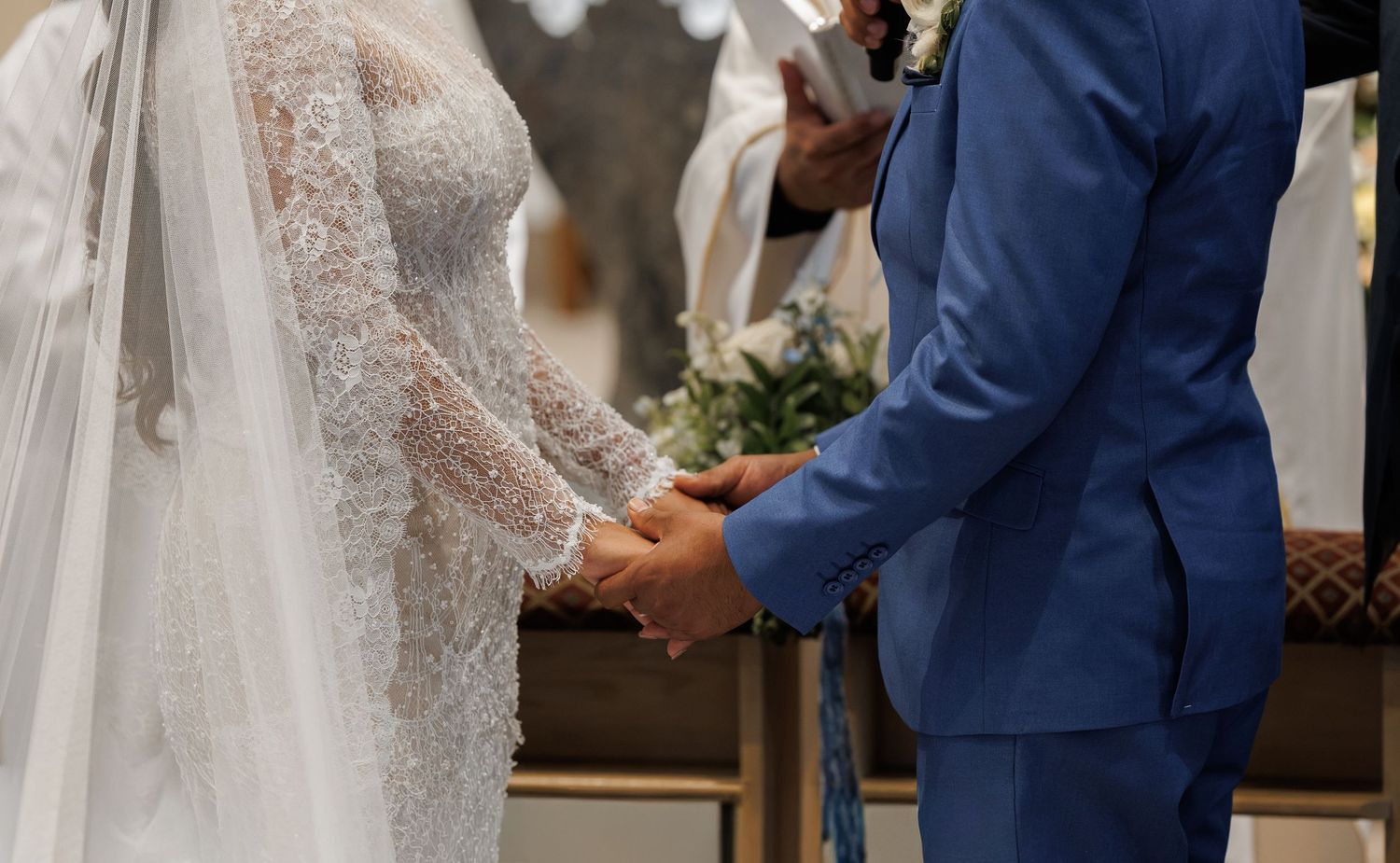 Bride in a lace wedding gown and veil holds hands with the groom in a blue suit during a church wedding ceremony.