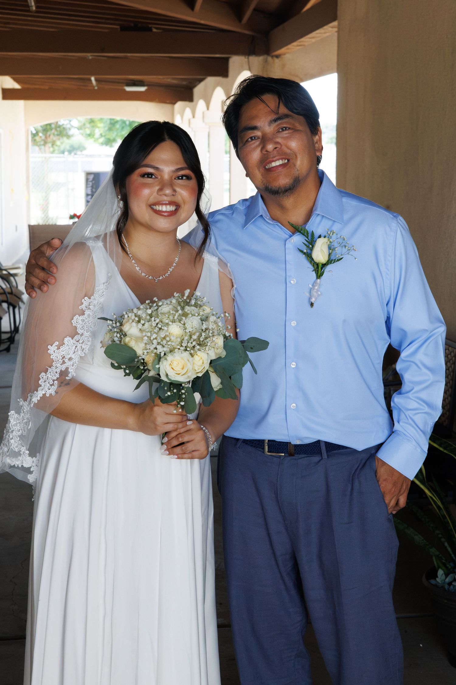 Smiling bride in white gown and veil holds bouquet beside groom in blue shirt with boutonniere at wedding.