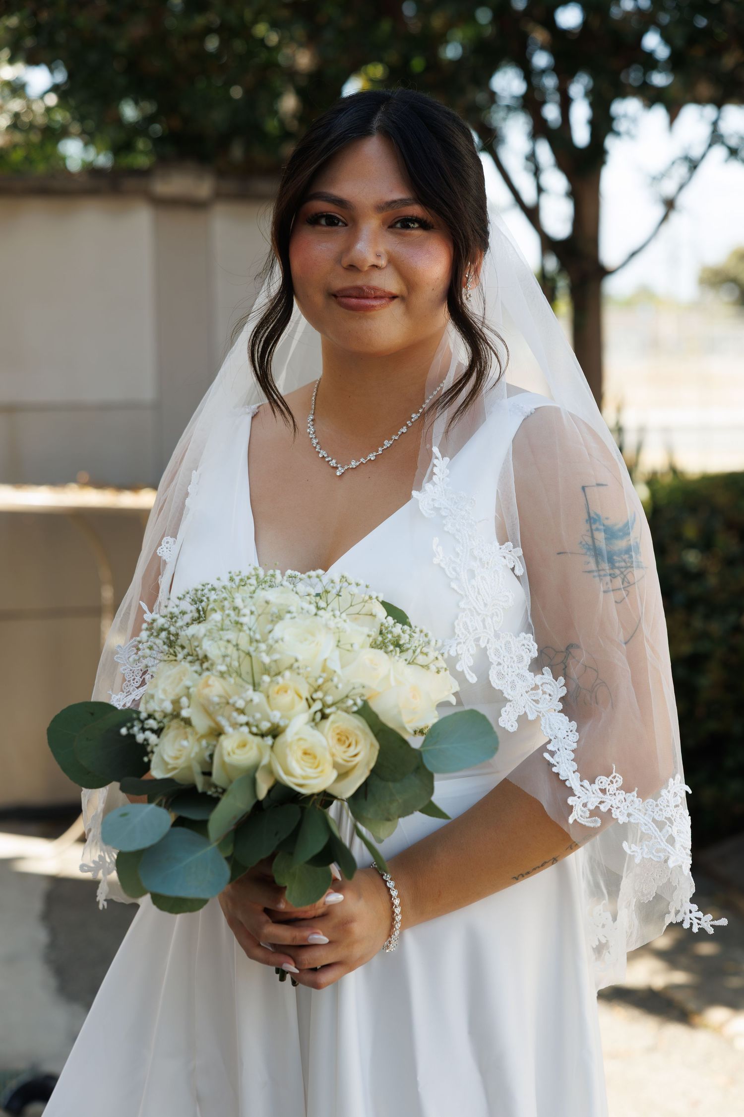Bride in white wedding dress and veil holding bouquet of white roses and eucalyptus, with pearl bracelet and diamond necklace.