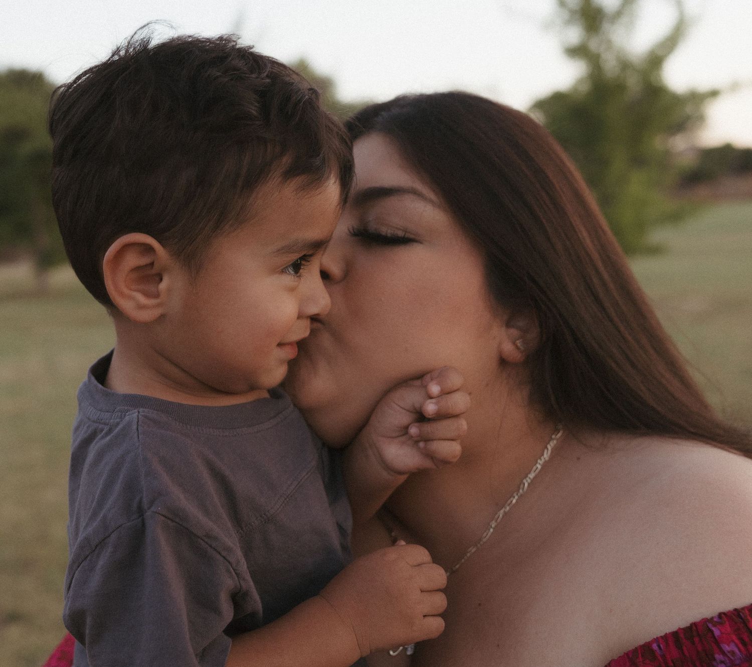 A loving mother kisses her young toddler son on the cheek outdoors in a warm, natural setting.