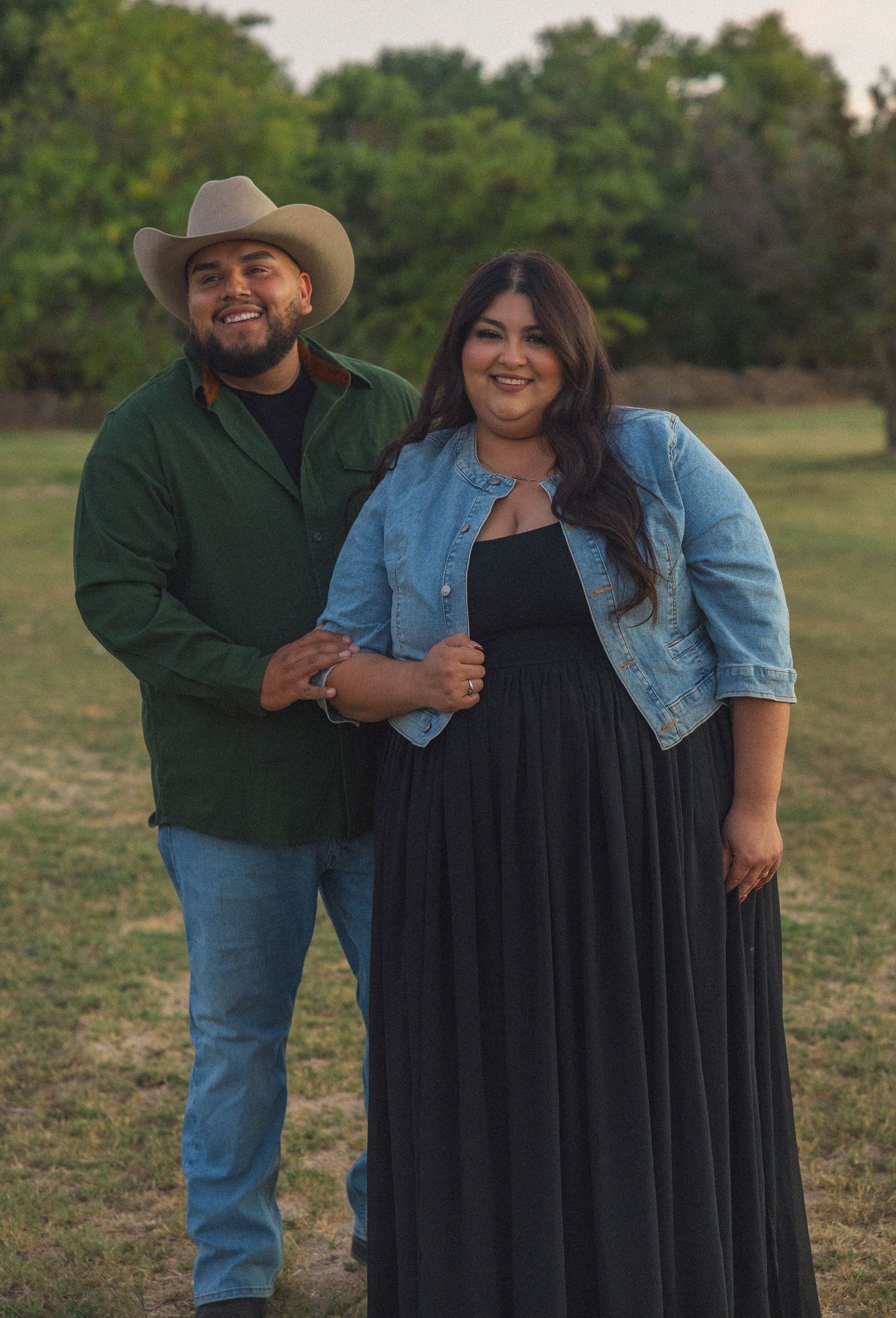 A couple poses outdoors in a green field, man in cowboy hat and green shirt, woman in black dress and denim jacket.