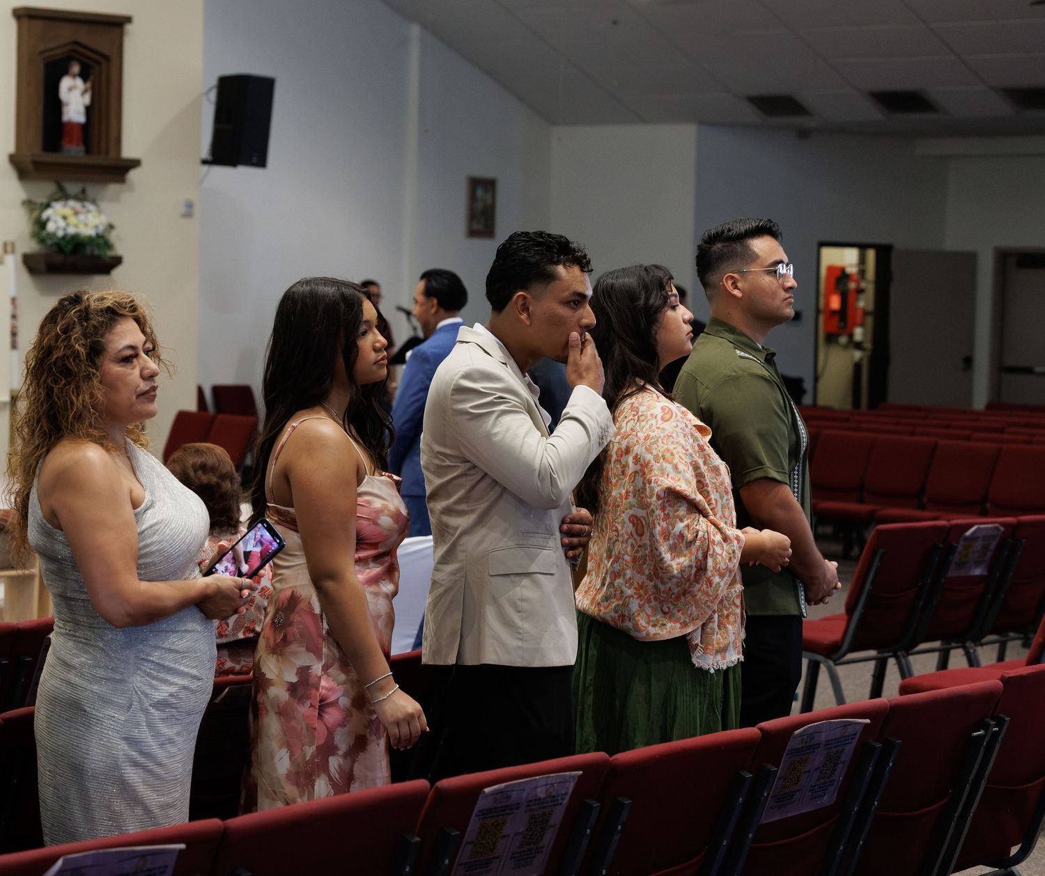 Congregation members stand in prayer between red church pews, with a religious statue shrine on the wall in the background.