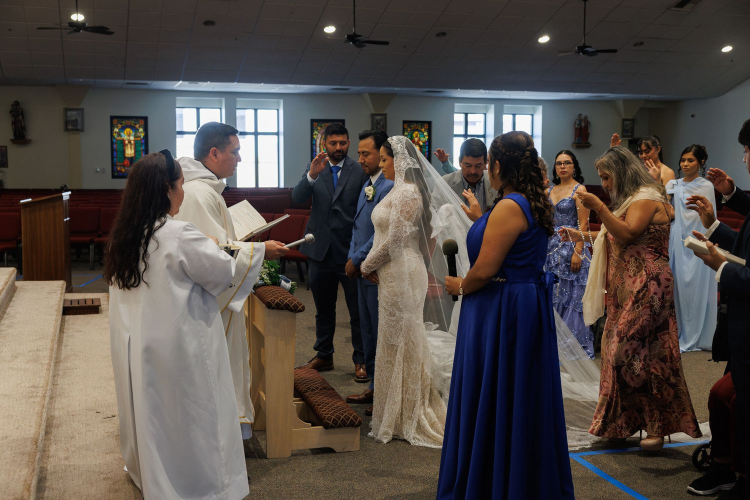 Bride in a white gown and veil stands with the groom before a priest in white robes during a church wedding ceremony as guests look on.