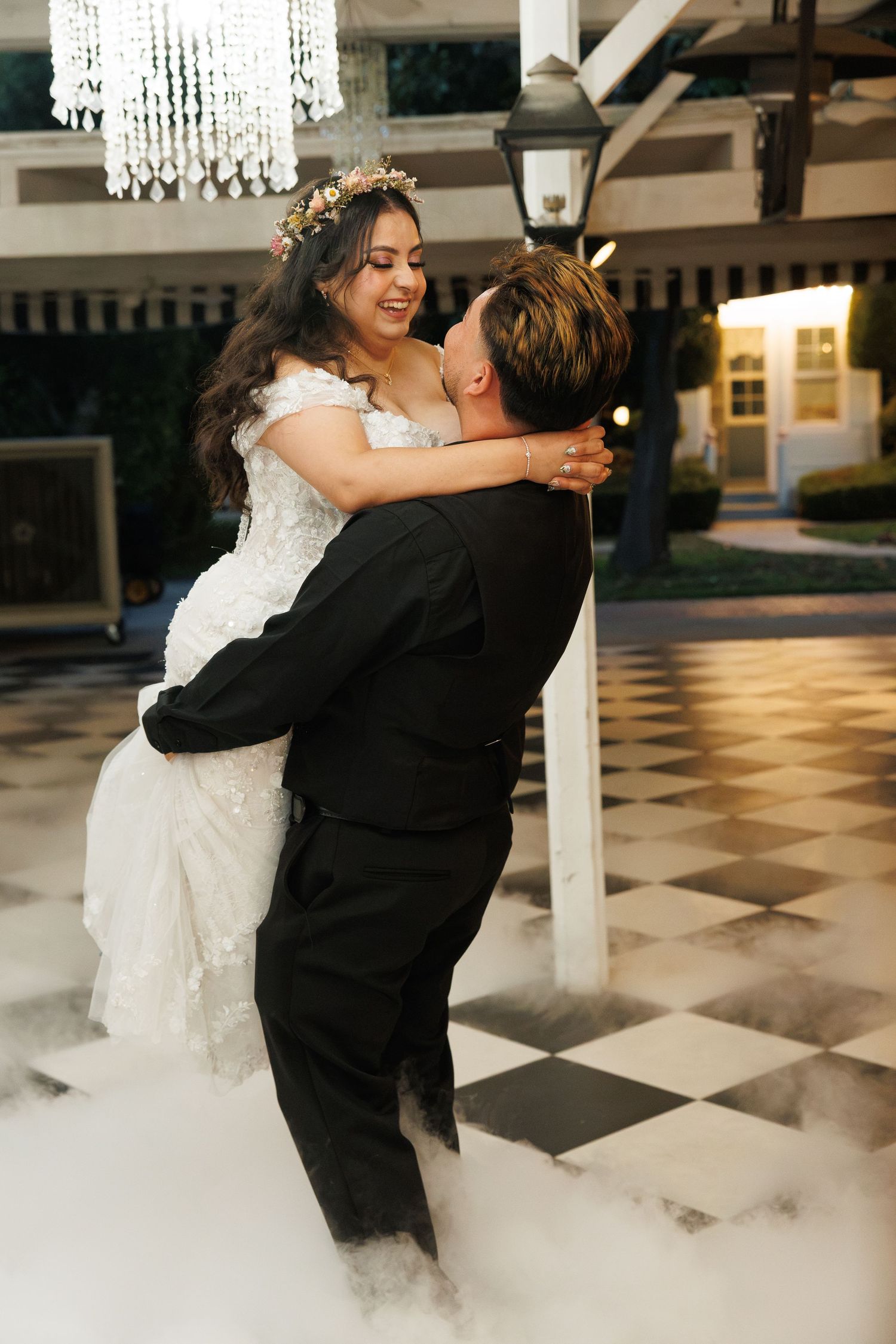 Groom lifts smiling bride in white gown on checkered dance floor surrounded by romantic fog at elegant wedding venue.
