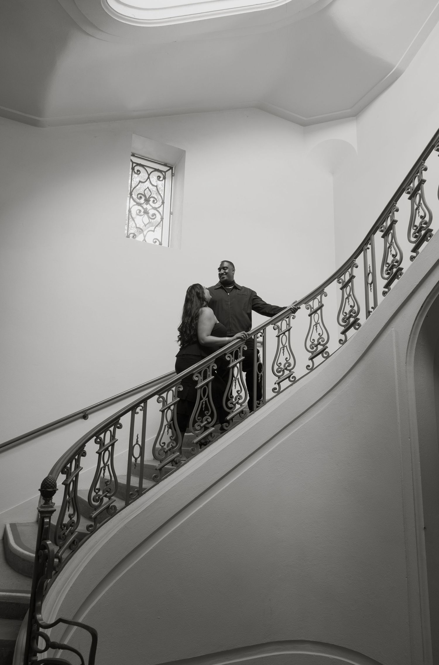 Couple posing on elegant curved staircase with ornate iron railing in black and white photography.