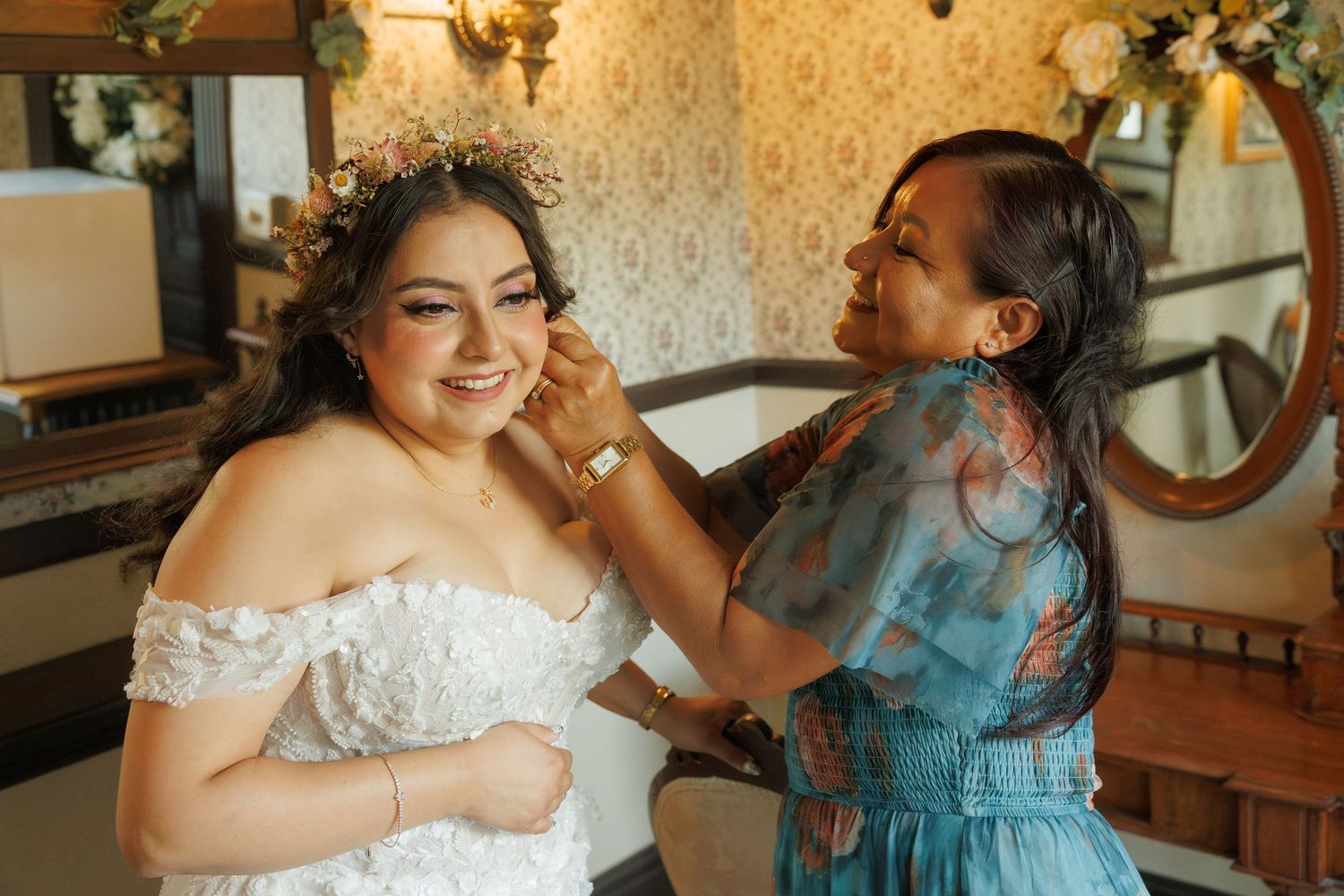 Bride in white lace off-shoulder gown smiles as woman in blue floral dress adjusts her earring in elegant room.
