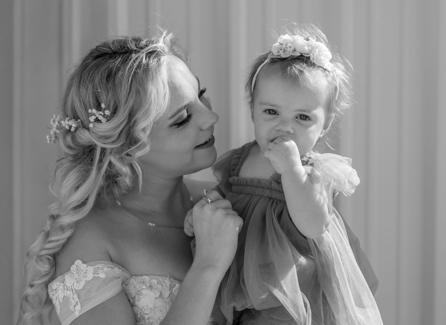Black and white photo of a bride holding a baby girl in a floral headband at a wedding ceremony.
