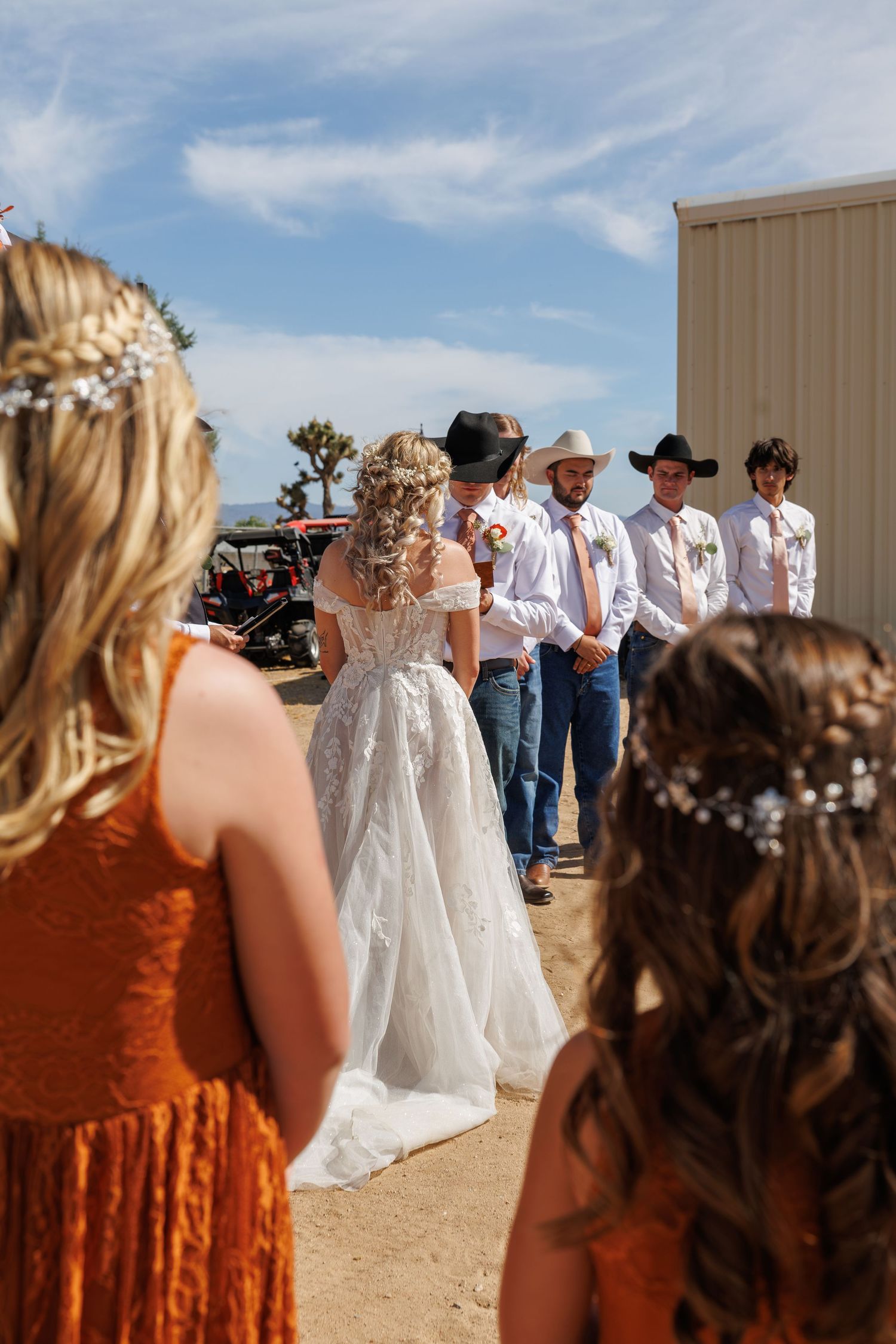 Bride in lace wedding gown meets groomsmen in cowboy hats and jeans at rustic outdoor western wedding ceremony.