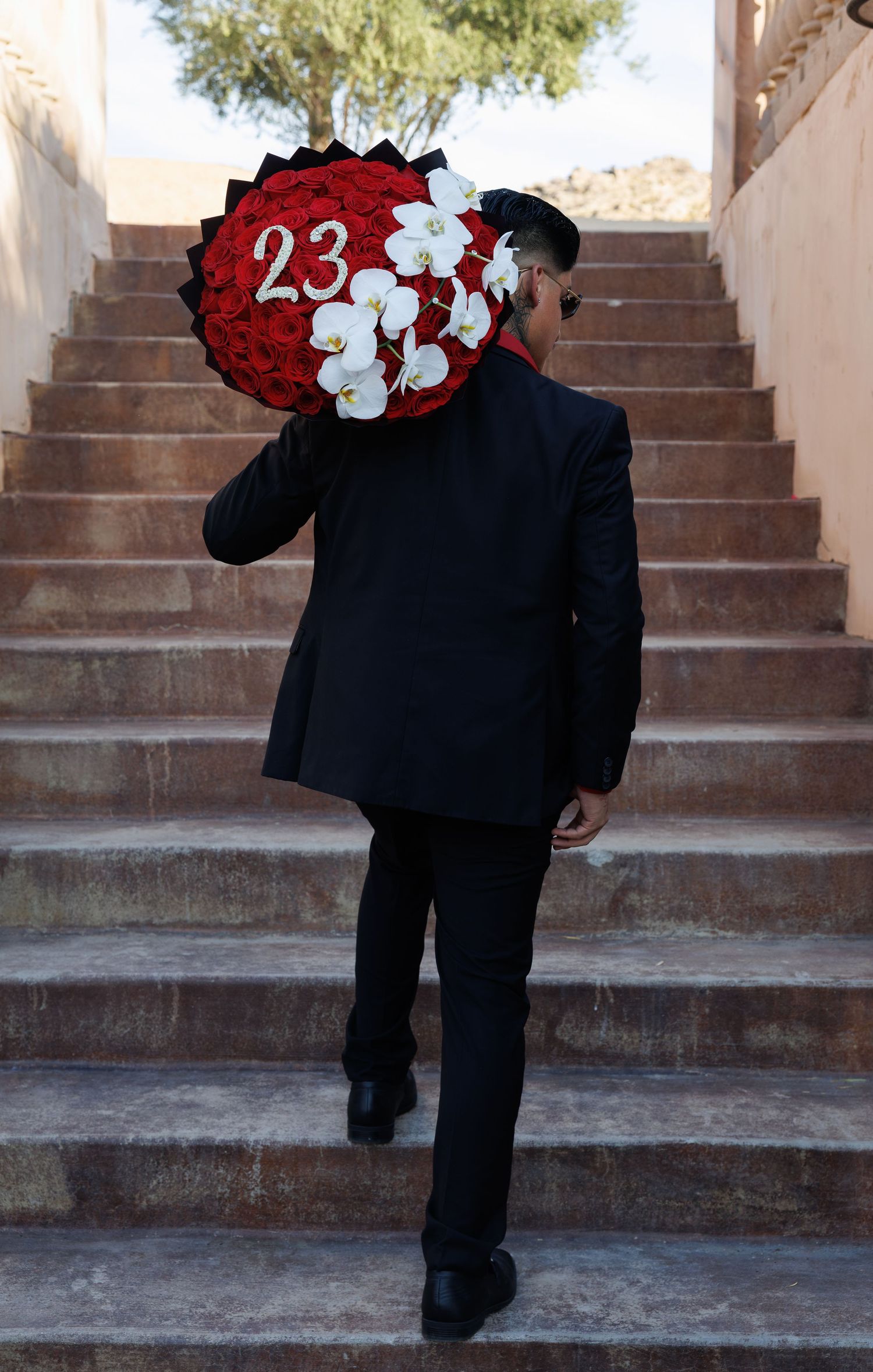 Man in black suit carrying large floral wreath with red roses and white orchids displaying number 23 up stone stairs.