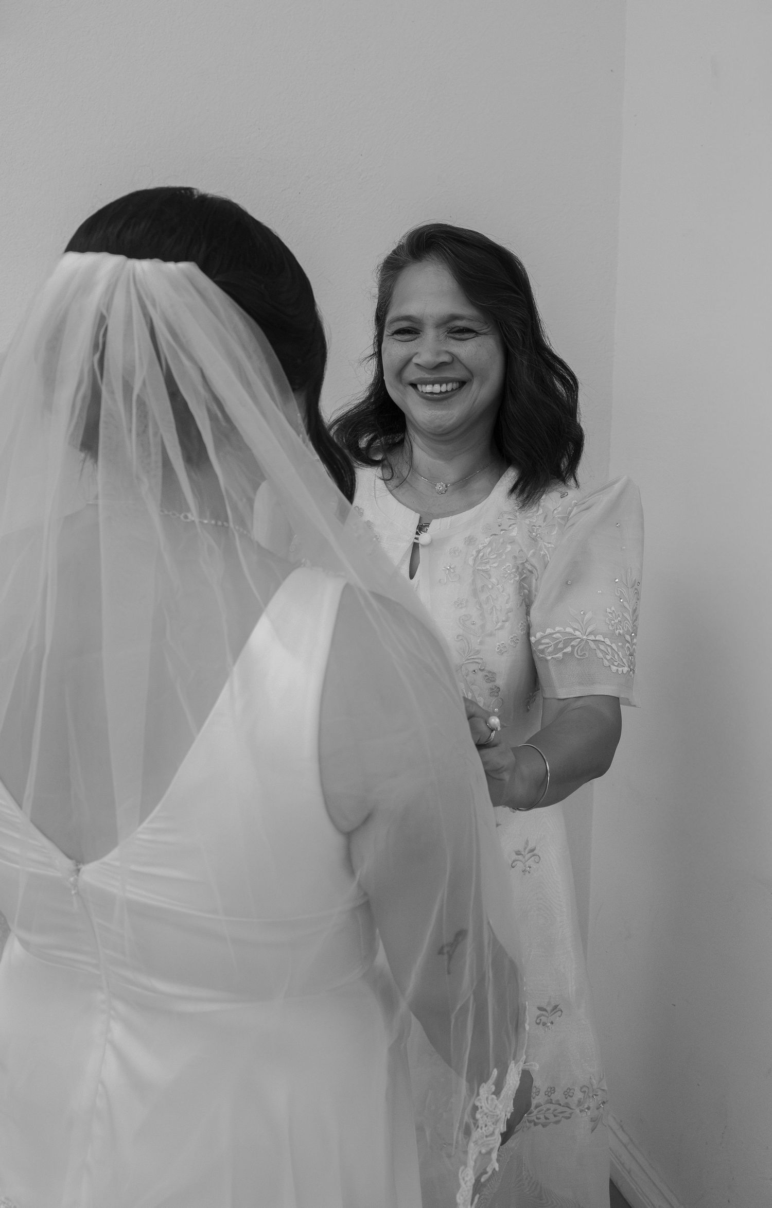 Black and white photo of a smiling woman in lace dress helping a bride in white gown and veil prepare for her wedding.