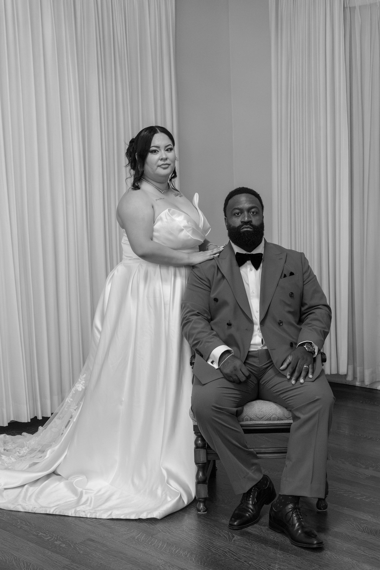 Black and white portrait of bride standing beside seated groom in tuxedo against white drapes.