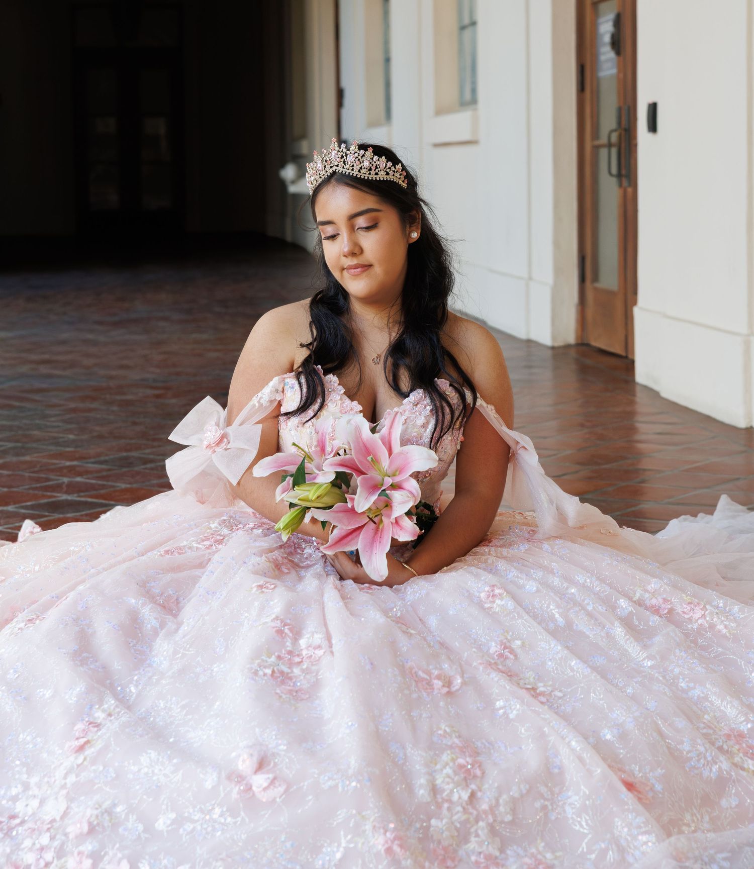 Young woman in pink floral ballgown with tiara holds lily bouquet during quinceañera portrait session.