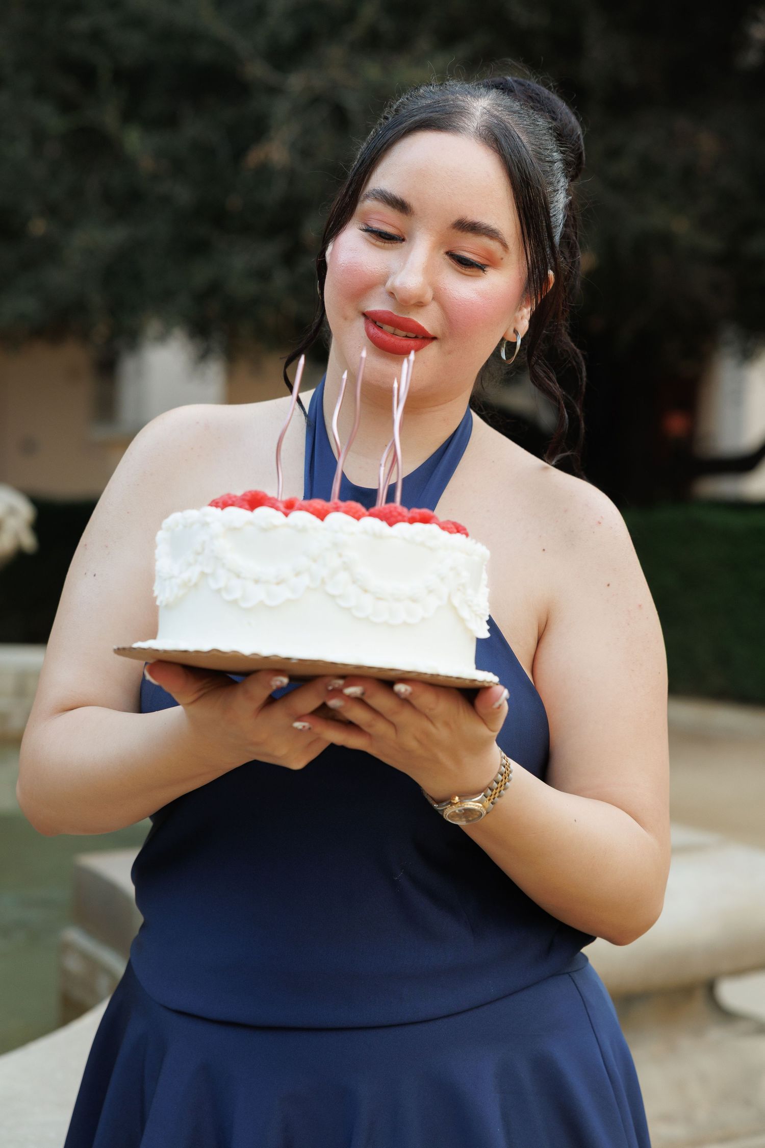 Smiling young woman in navy halter dress holding white raspberry birthday cake with candles in garden.