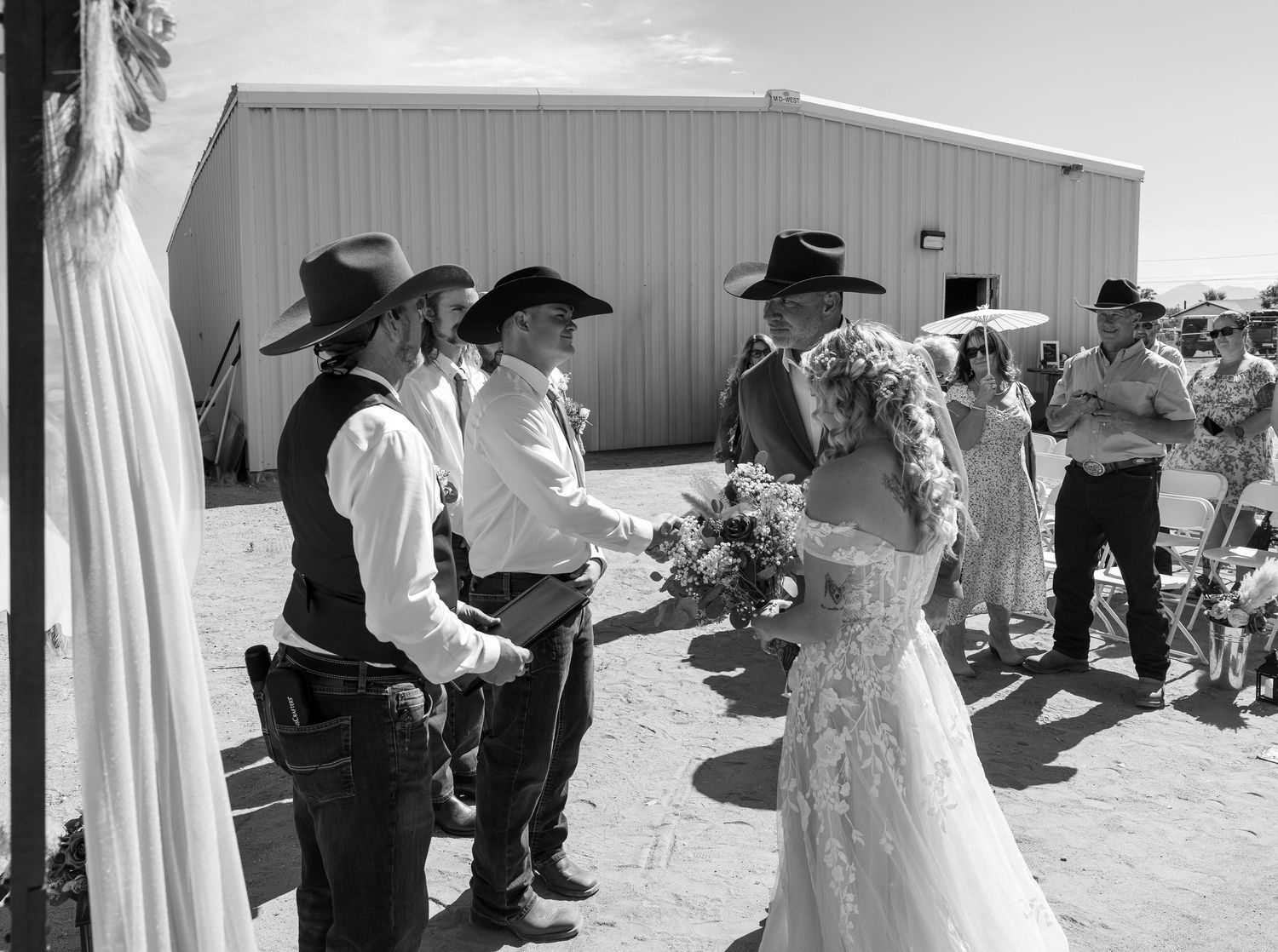 Black and white photo of a bride in a lace gown greeting cowboy-hat-wearing guests at an outdoor western wedding.