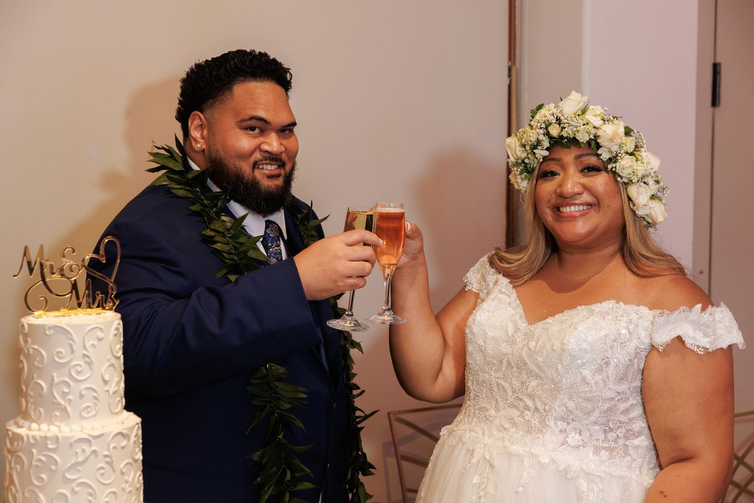Bride and groom toasting with champagne glasses beside wedding cake, bride wearing floral crown and groom in blue suit with lei.