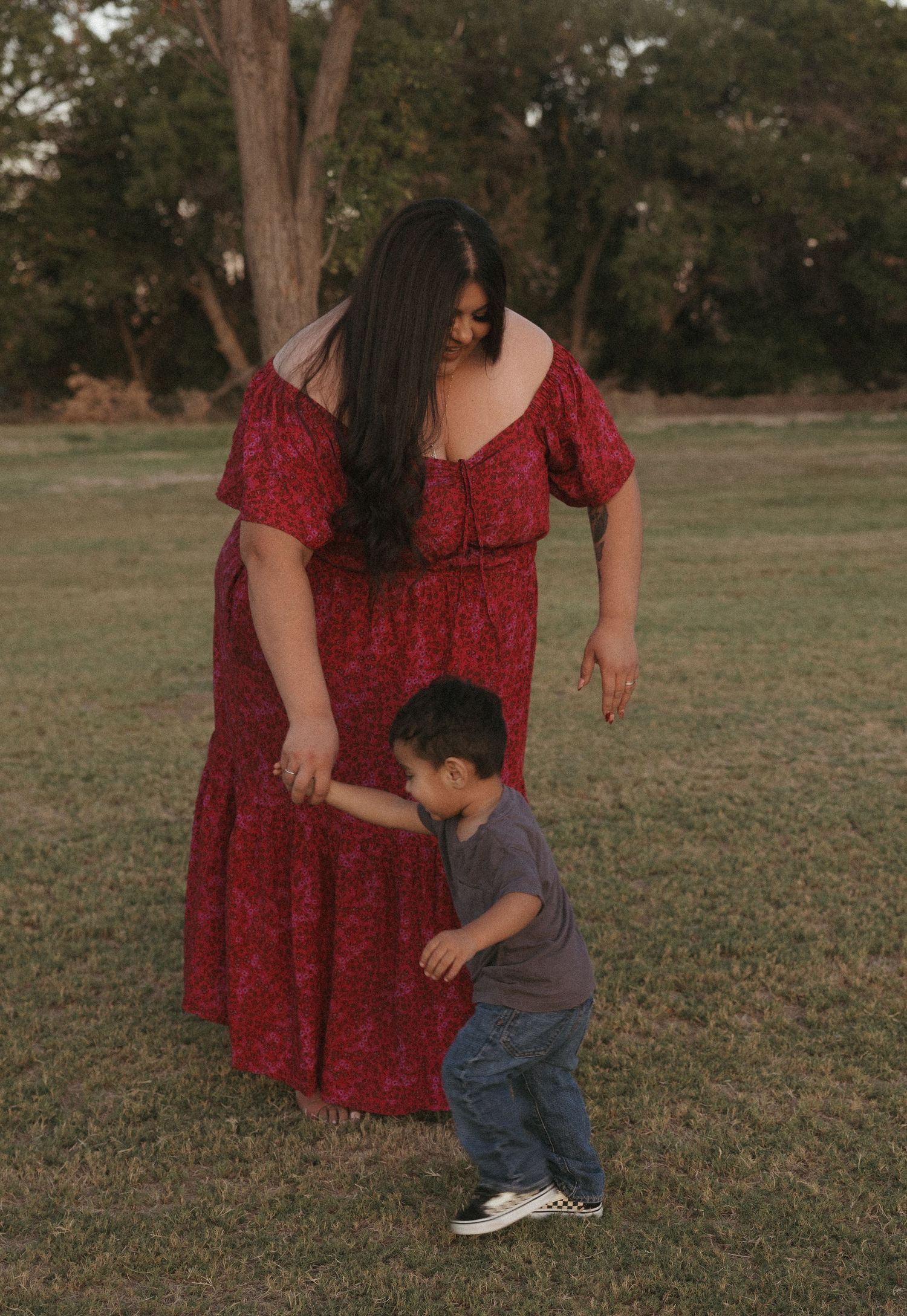 A mother in a red floral dress holds hands with her toddler son on a grassy park lawn at golden hour.