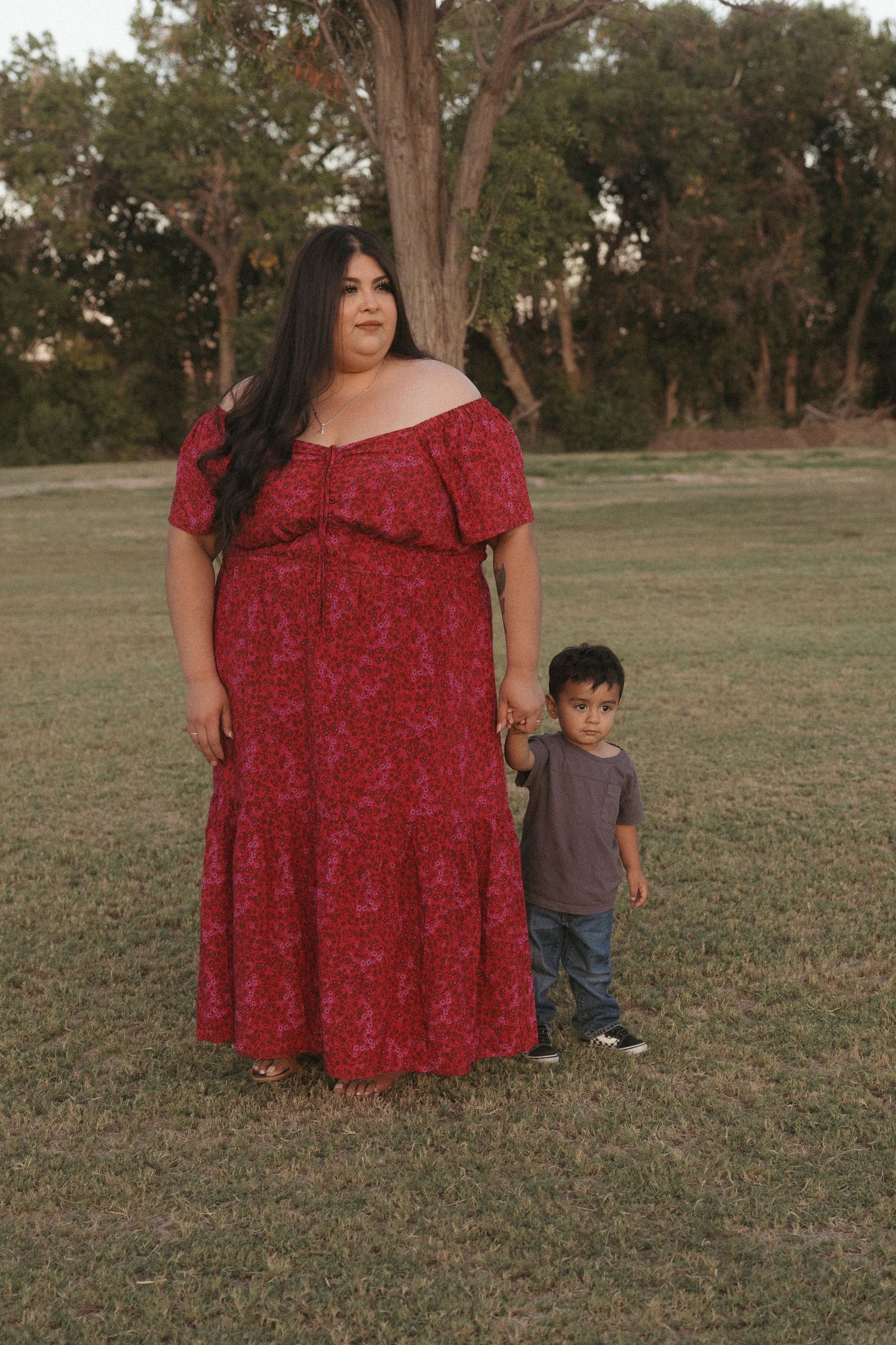 Woman in red floral off-shoulder dress holds hands with young boy in park setting near large tree.