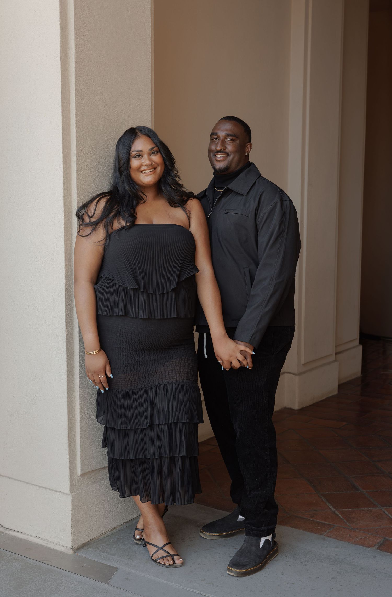 A couple dressed in matching black outfits pose together in an elegant hallway, smiling and holding hands.