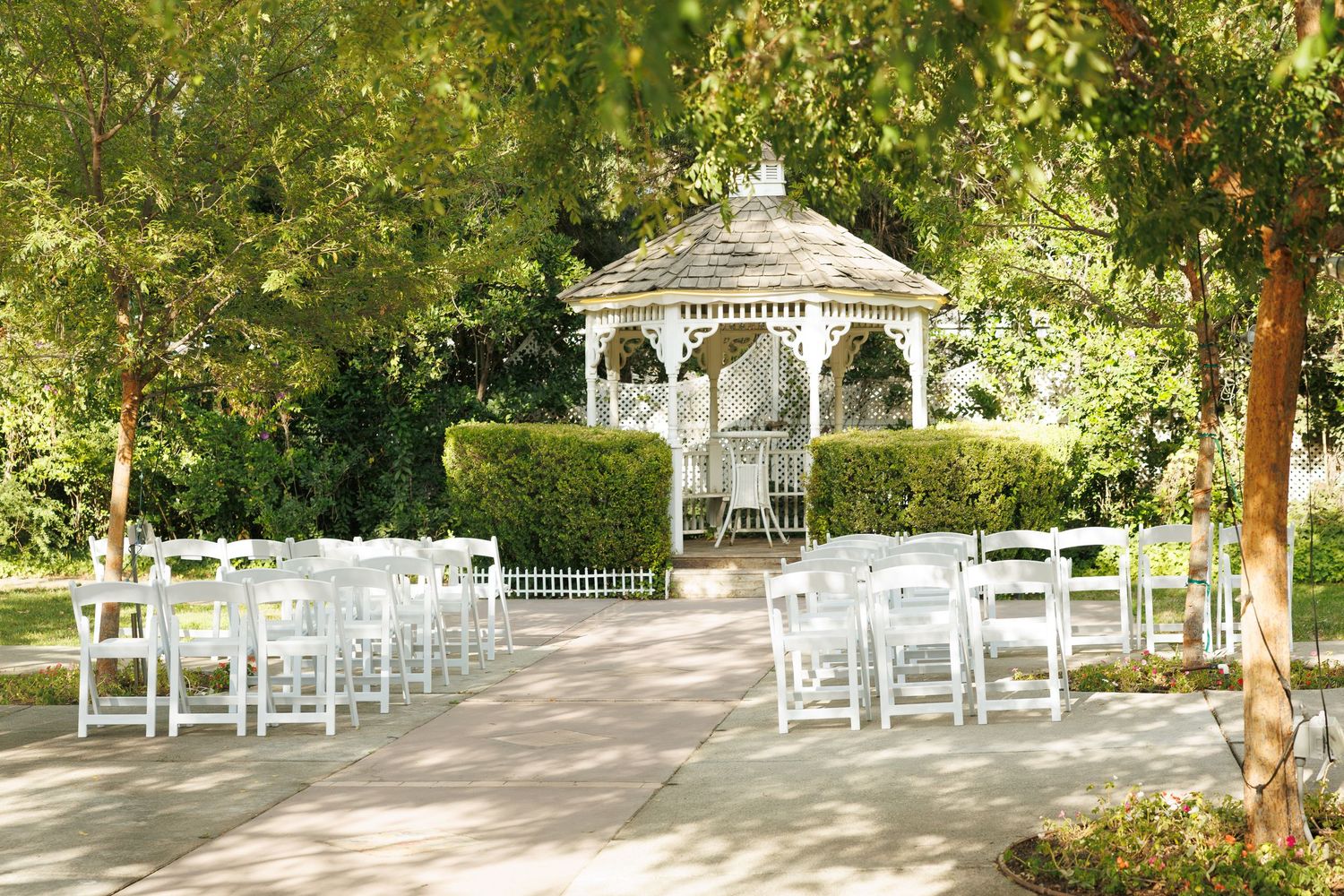 White chairs arranged before a charming white gazebo surrounded by lush green trees for an outdoor wedding ceremony.