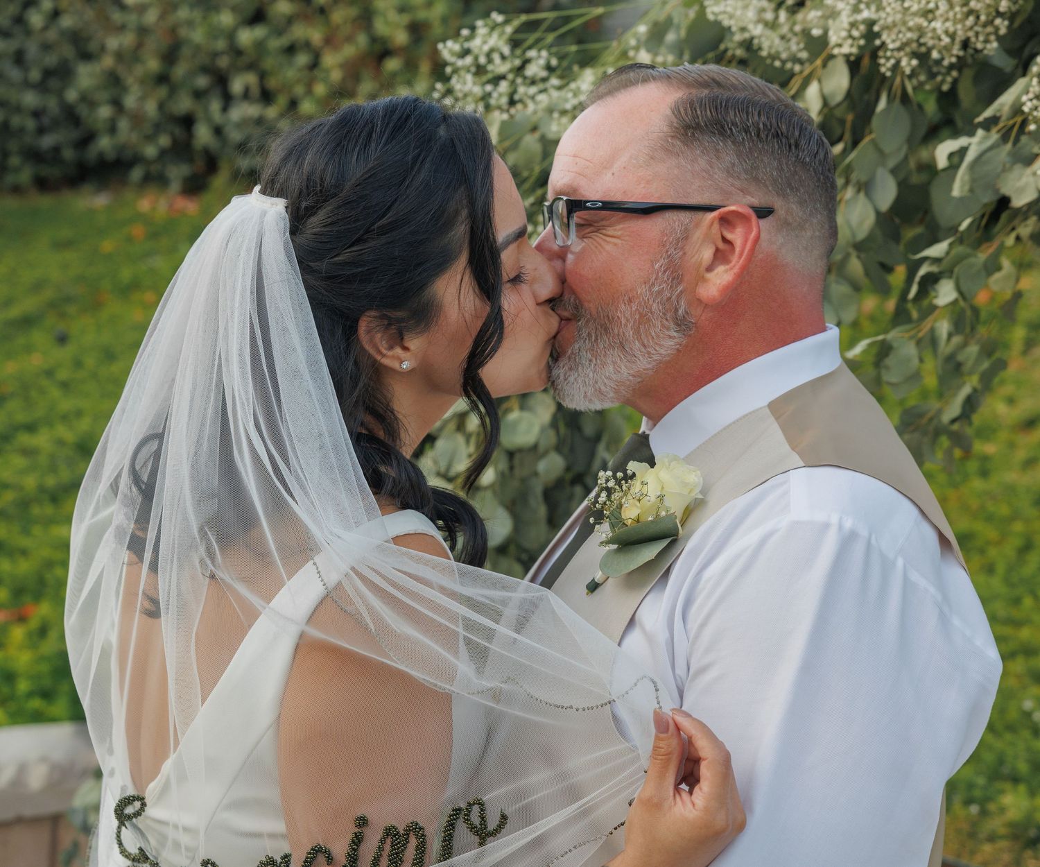 Bride and groom share a romantic first kiss at outdoor wedding ceremony, bride wearing veil and white gown.