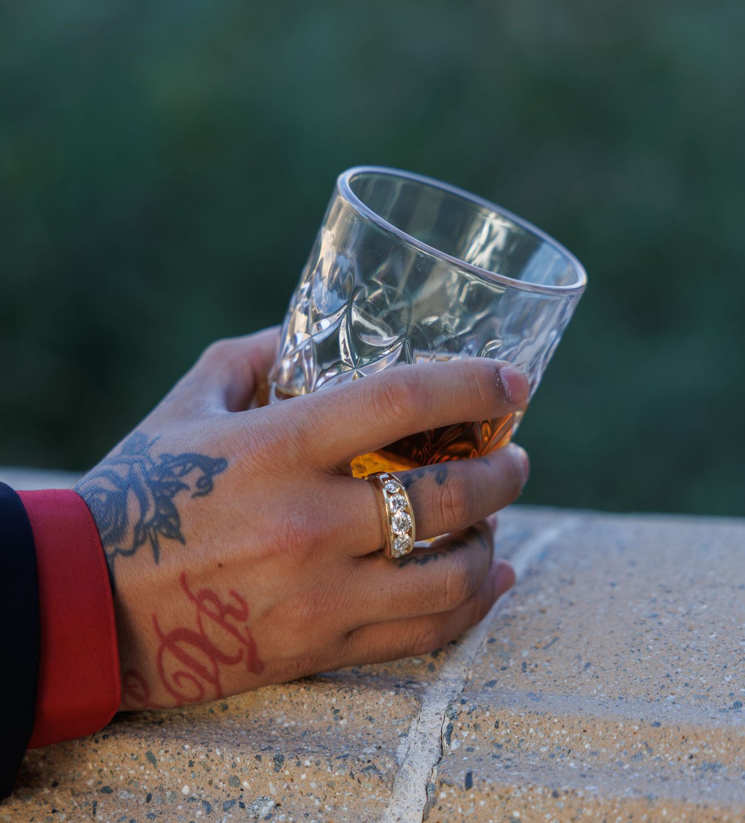 Tattooed hand with gold ring holding a crystal whiskey glass on a brick ledge outdoors.