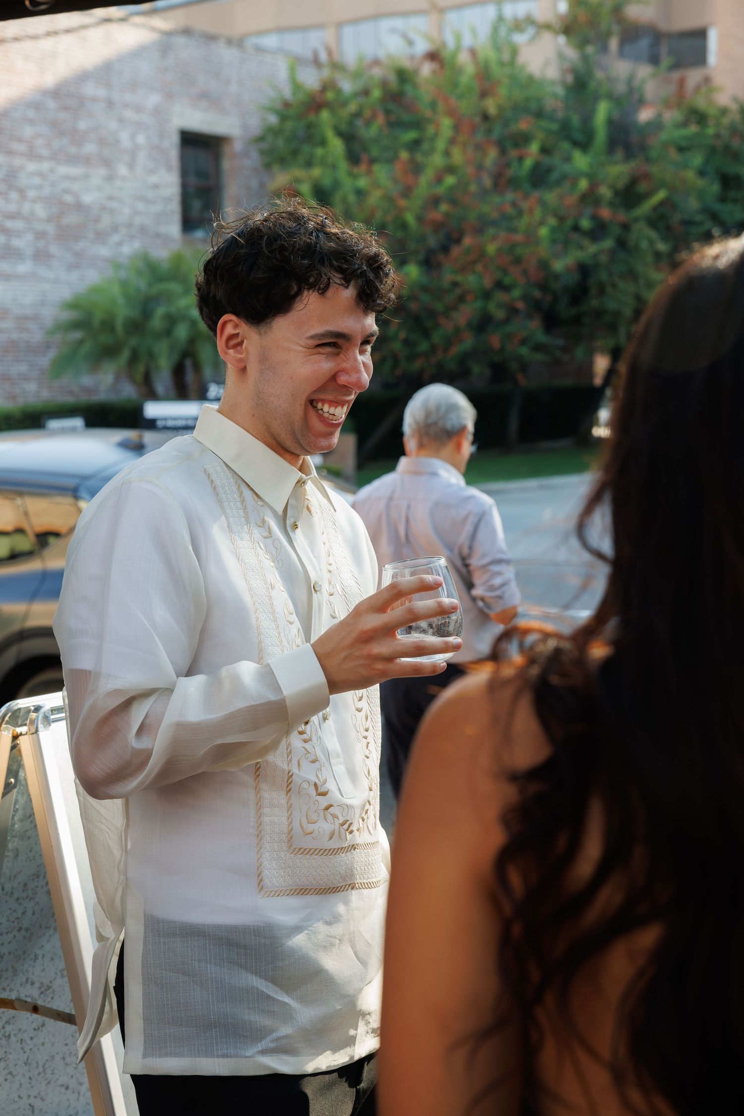 Smiling young man in traditional Filipino barong tagalog holding a drink glass at an outdoor wedding reception.