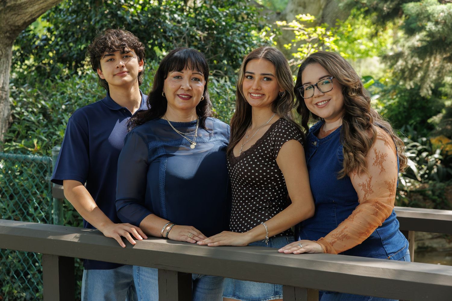 Four smiling family members pose together on a wooden bridge surrounded by lush green trees outdoors.