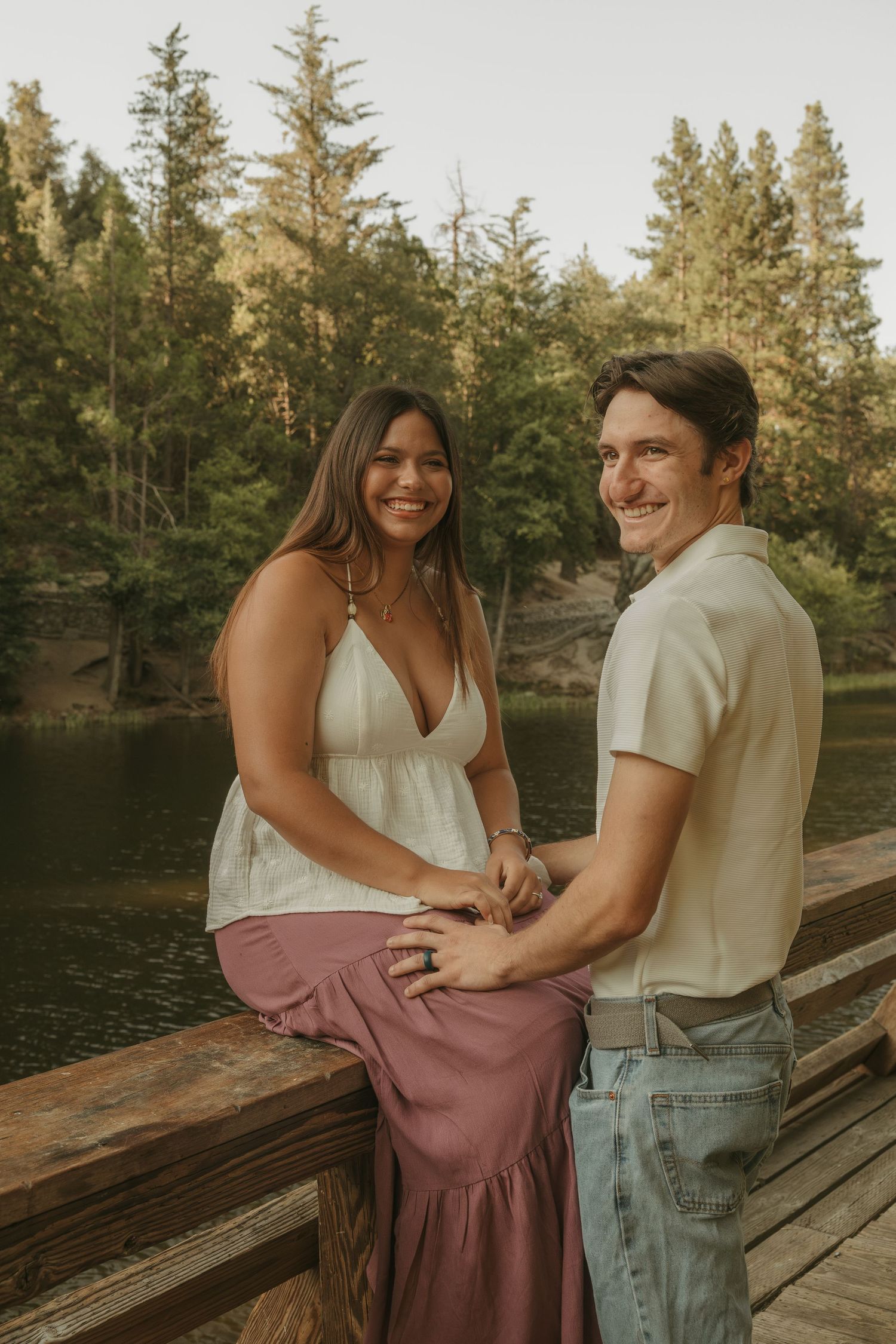 Smiling couple holding hands on a wooden bridge by a lake surrounded by tall evergreen trees.