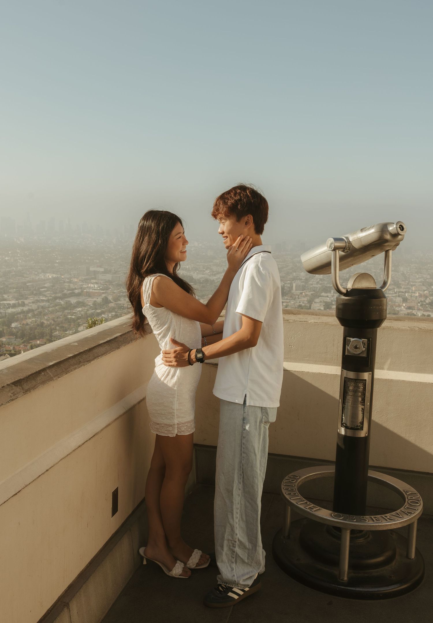 A couple embraces romantically on a scenic overlook with a coin-operated telescope and city skyline backdrop.