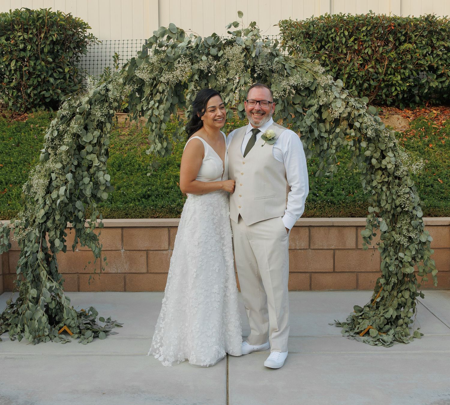 Bride and groom smiling under a lush greenery arch at an outdoor wedding ceremony.