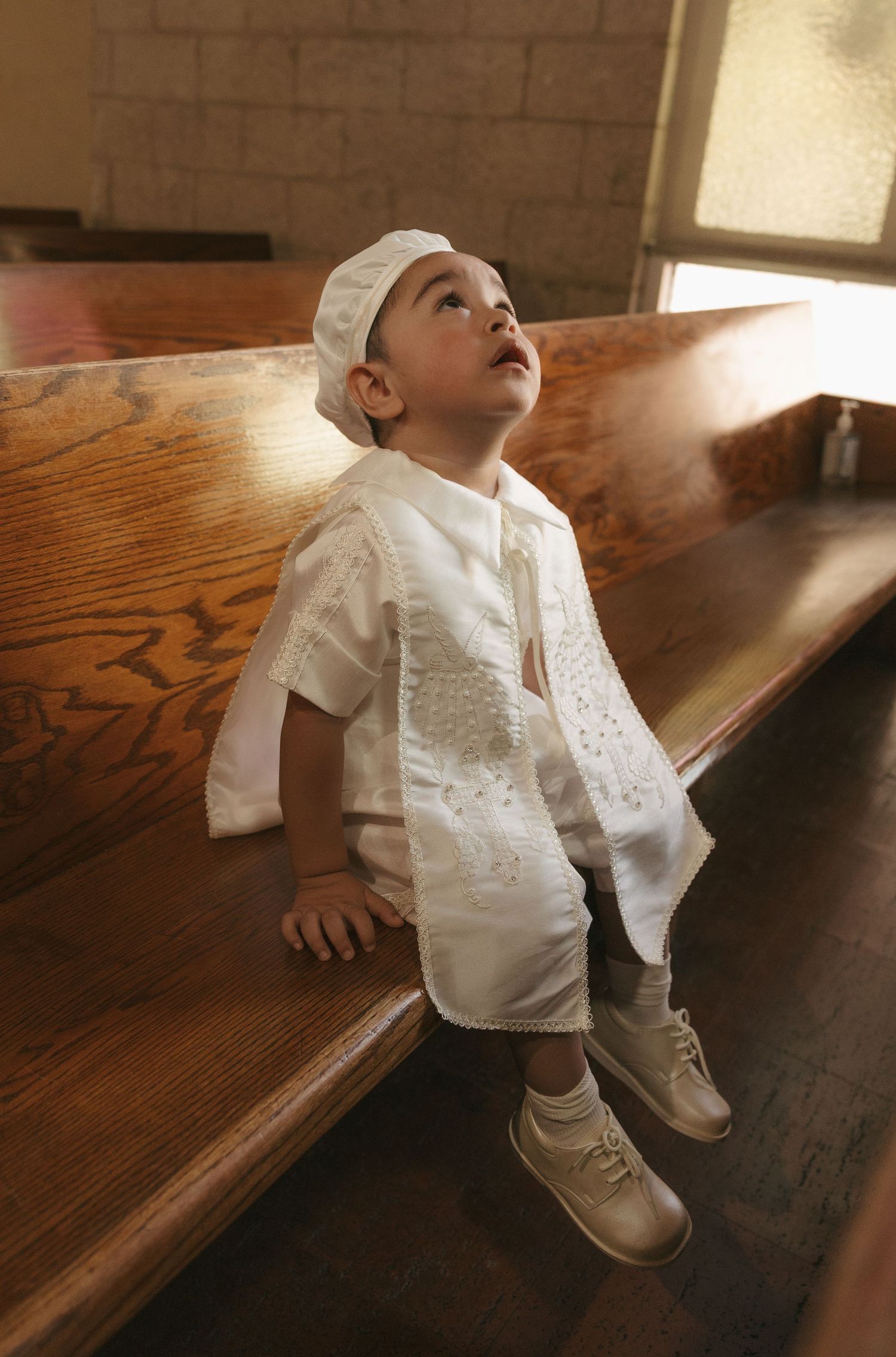Toddler in white christening outfit sitting on church pew, looking up with wonder.