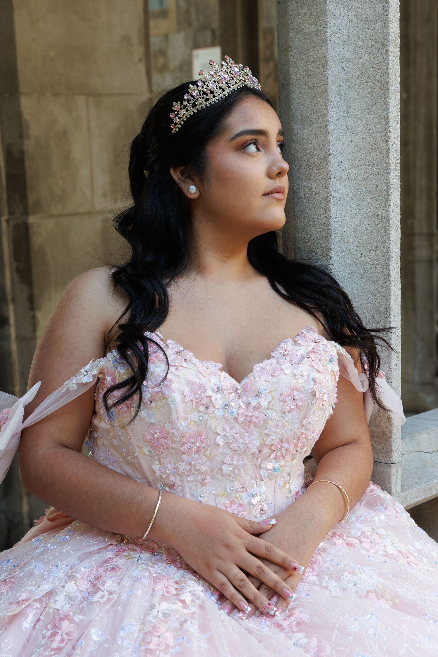 Young Latina in pink floral quinceañera ball gown and tiara sits gracefully against stone column.