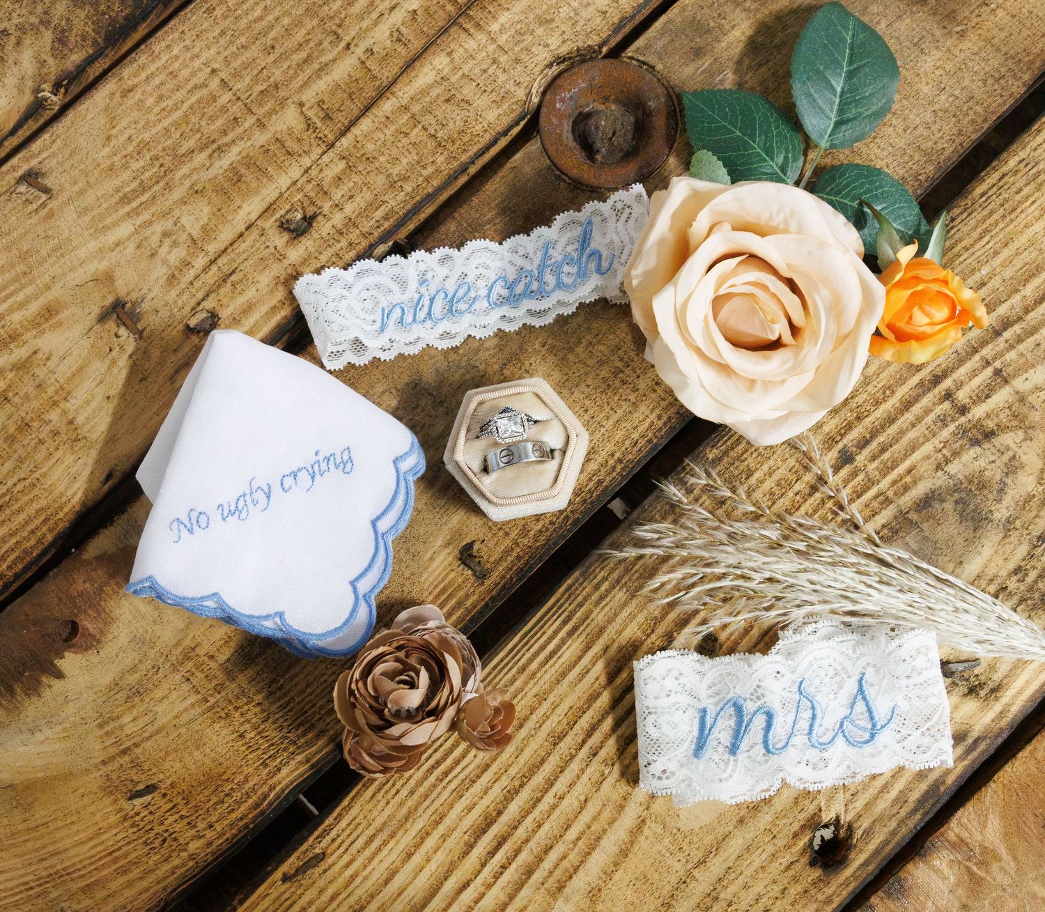 Wedding flat lay with embroidered handkerchief, ring box, lace garter, and floral decorations on wood.