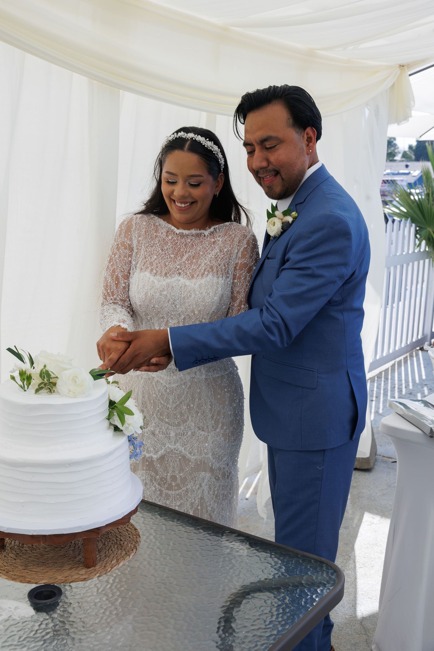 Smiling bride in a lace dress and groom in a blue suit cut their white tiered wedding cake together under a white canopy.