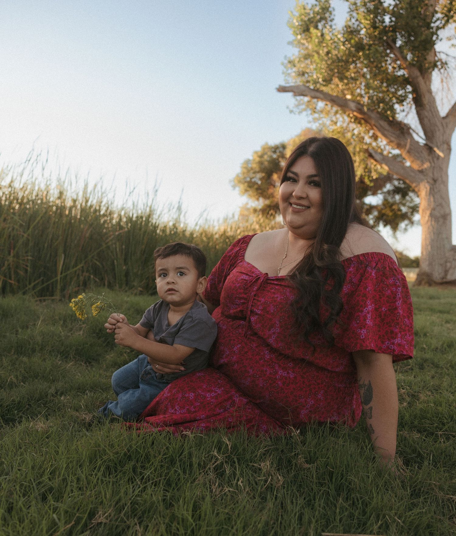 A young woman in a red floral dress sits on green grass with a toddler boy holding flowers, outdoors near trees.