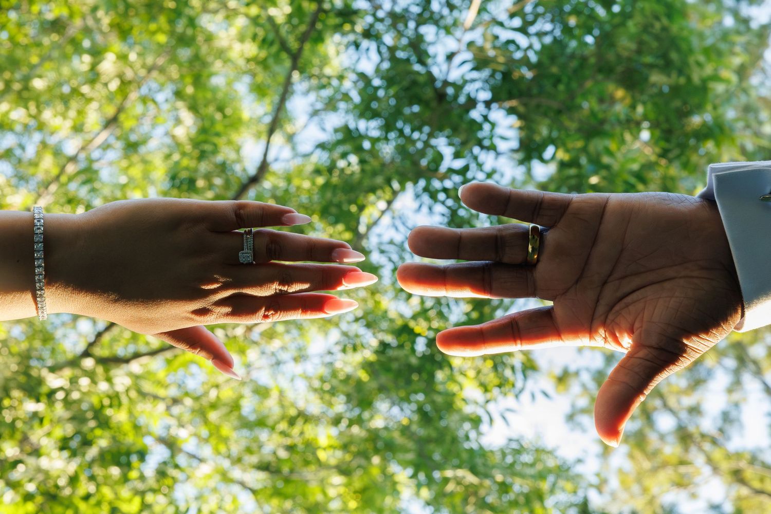 Couple displaying wedding rings outdoors with green trees in background on their wedding day.
