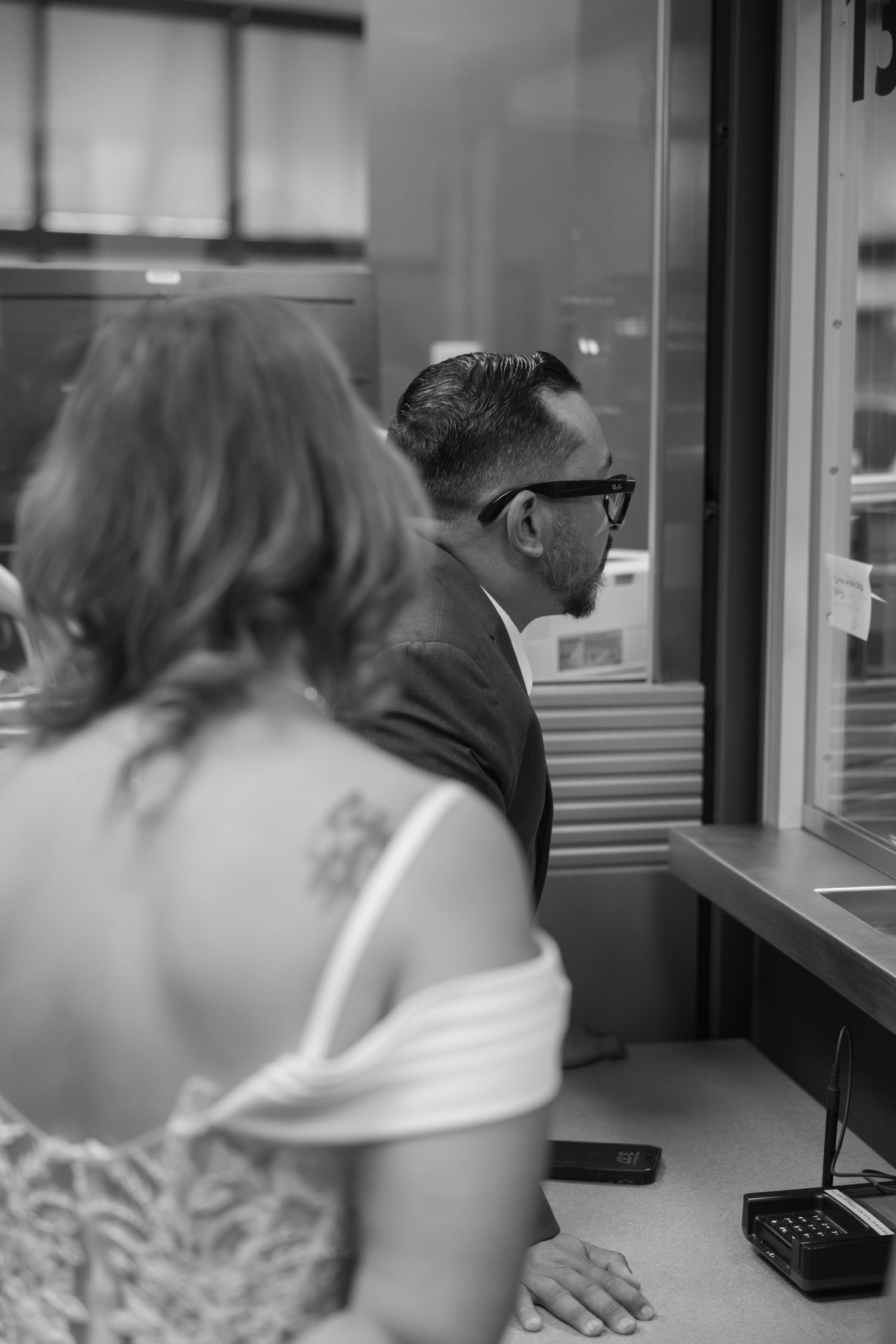 Black and white photo of a couple at a service window, woman with floral tattoo in lace dress, man in suit wearing glasses.