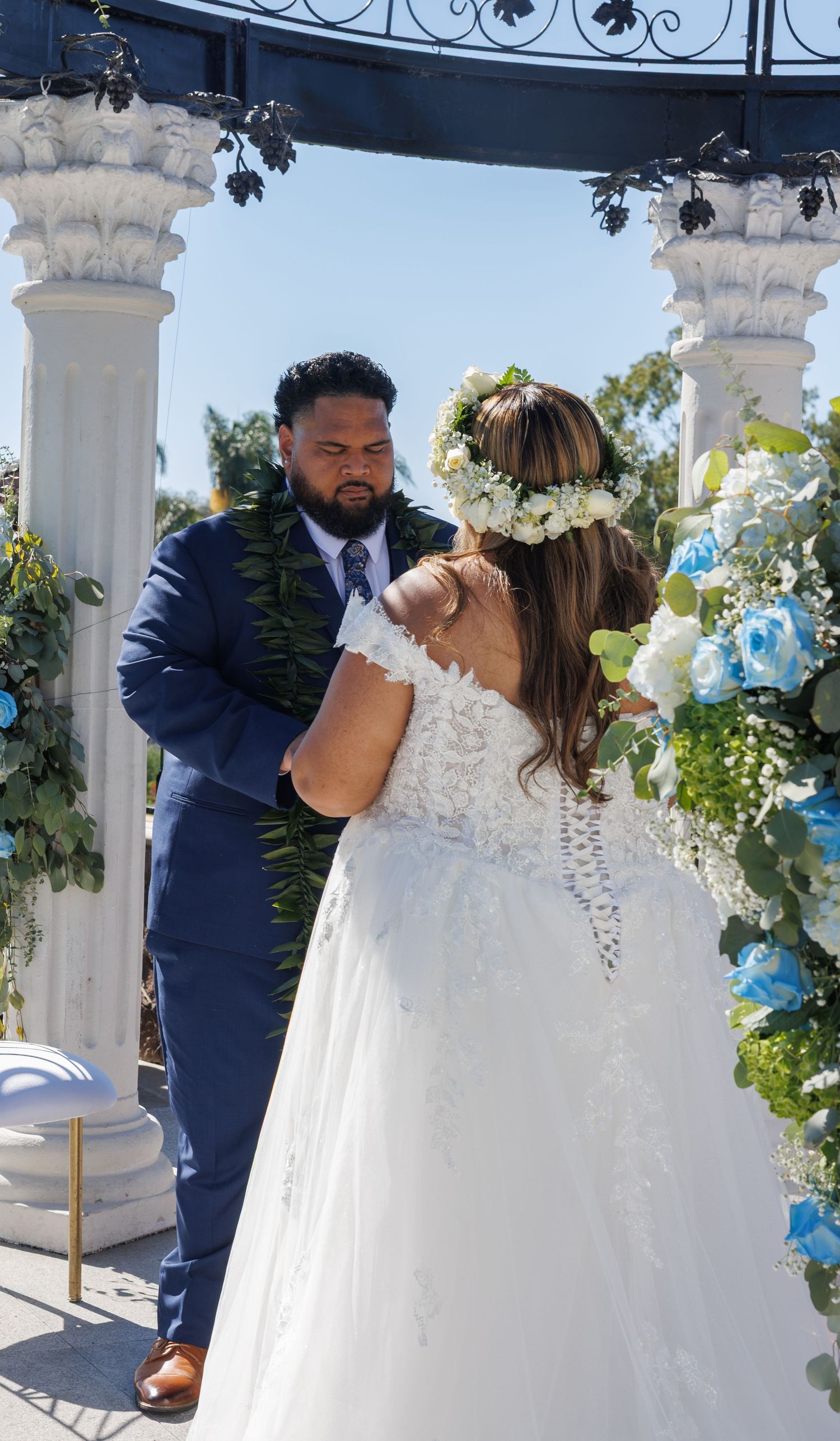 Bride in white lace gown with floral crown and groom in navy suit share a tender moment at an outdoor wedding ceremony.