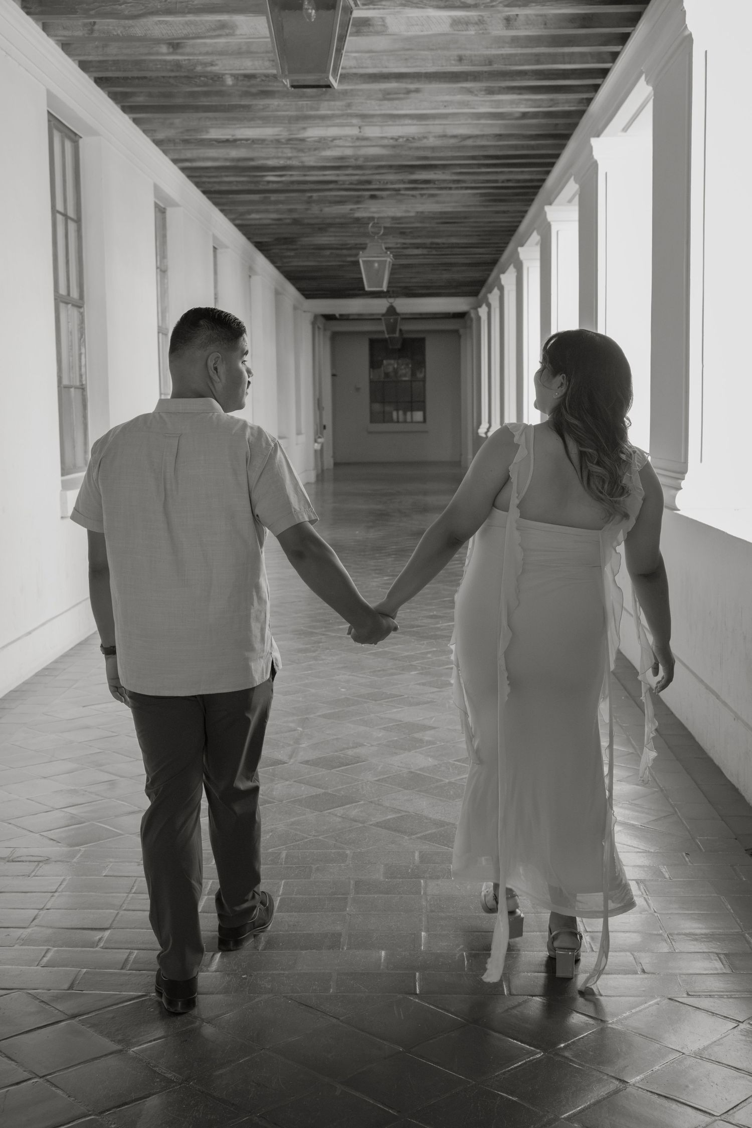Couple holding hands walking down elegant corridor, black and white maternity photo showing love and connection.