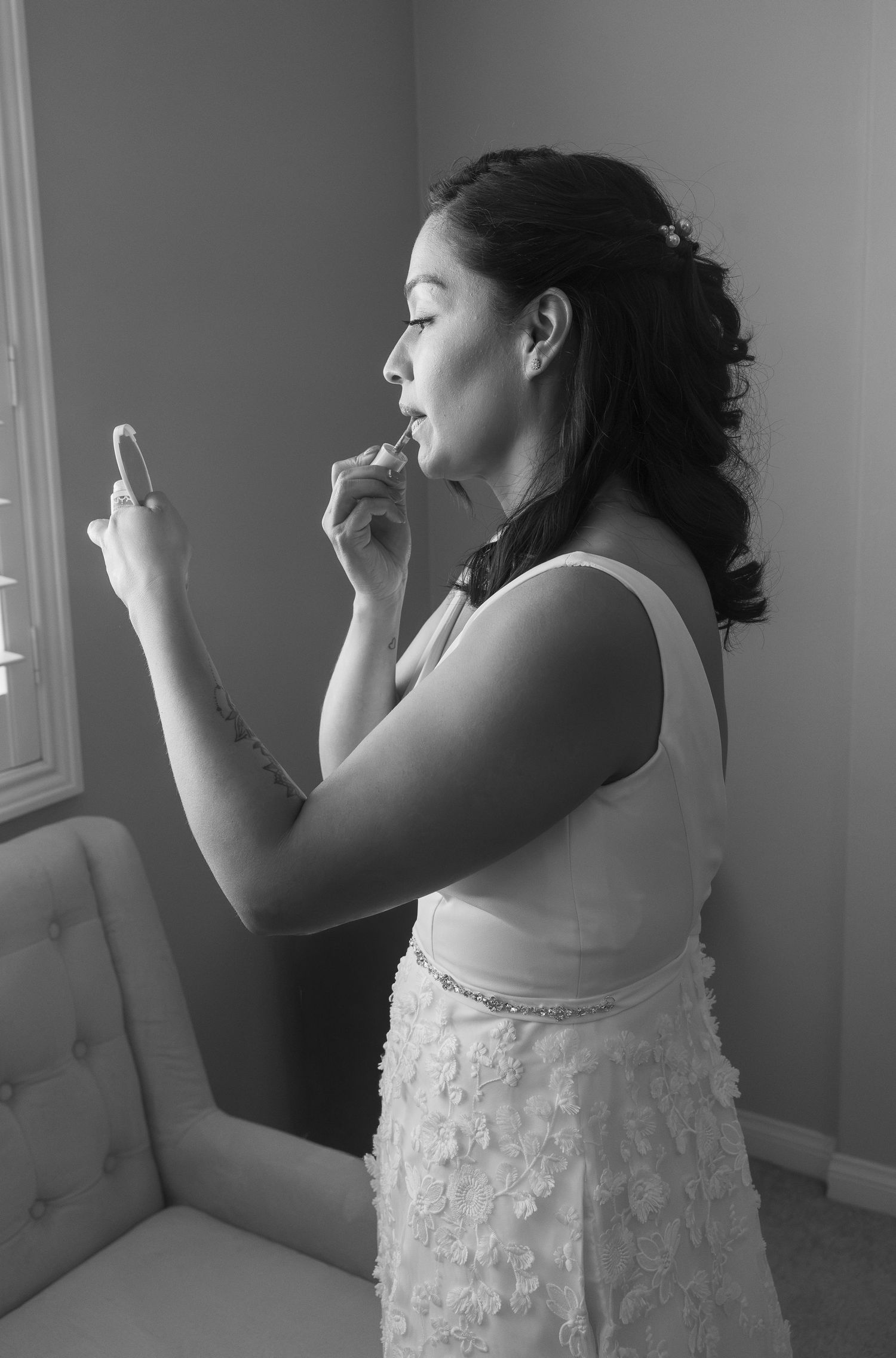 Bride in floral wedding gown applies lipstick using compact mirror near window in black and white photo.