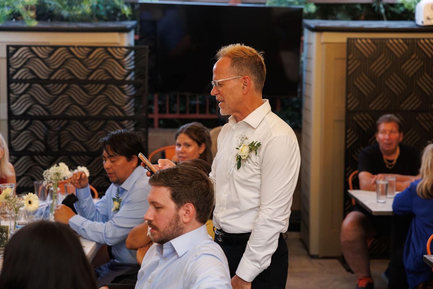 Man in white shirt with boutonniere stands at wedding reception while guests sit at decorated tables.