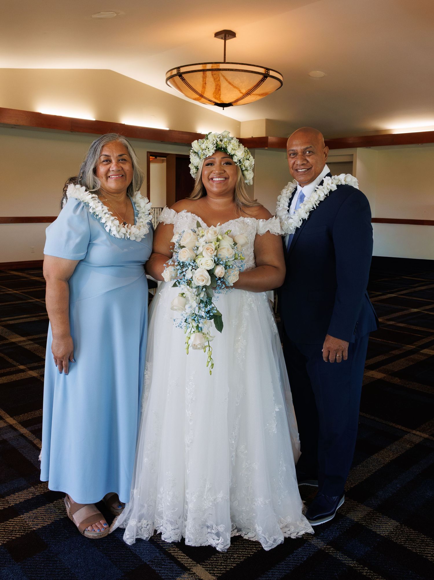 Bride in white lace gown with floral crown holds bouquet, flanked by two guests wearing floral leis in blue attire.