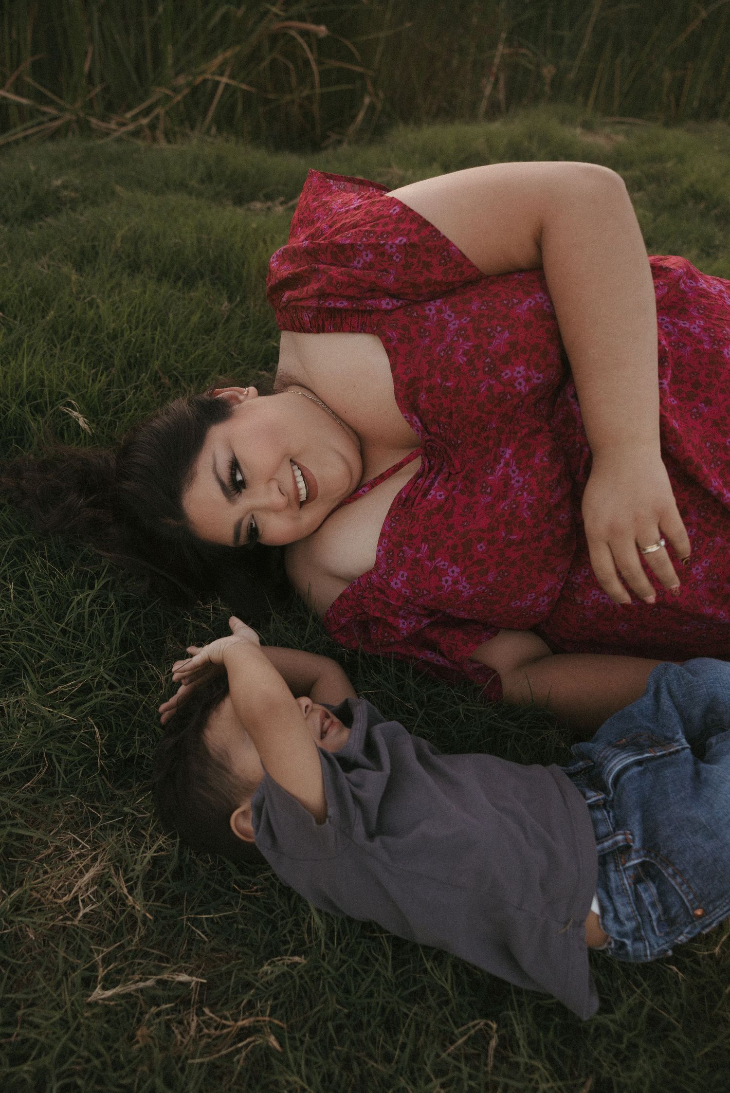 A woman in a red floral dress lies on grass smiling at a young toddler boy laying beside her outdoors.