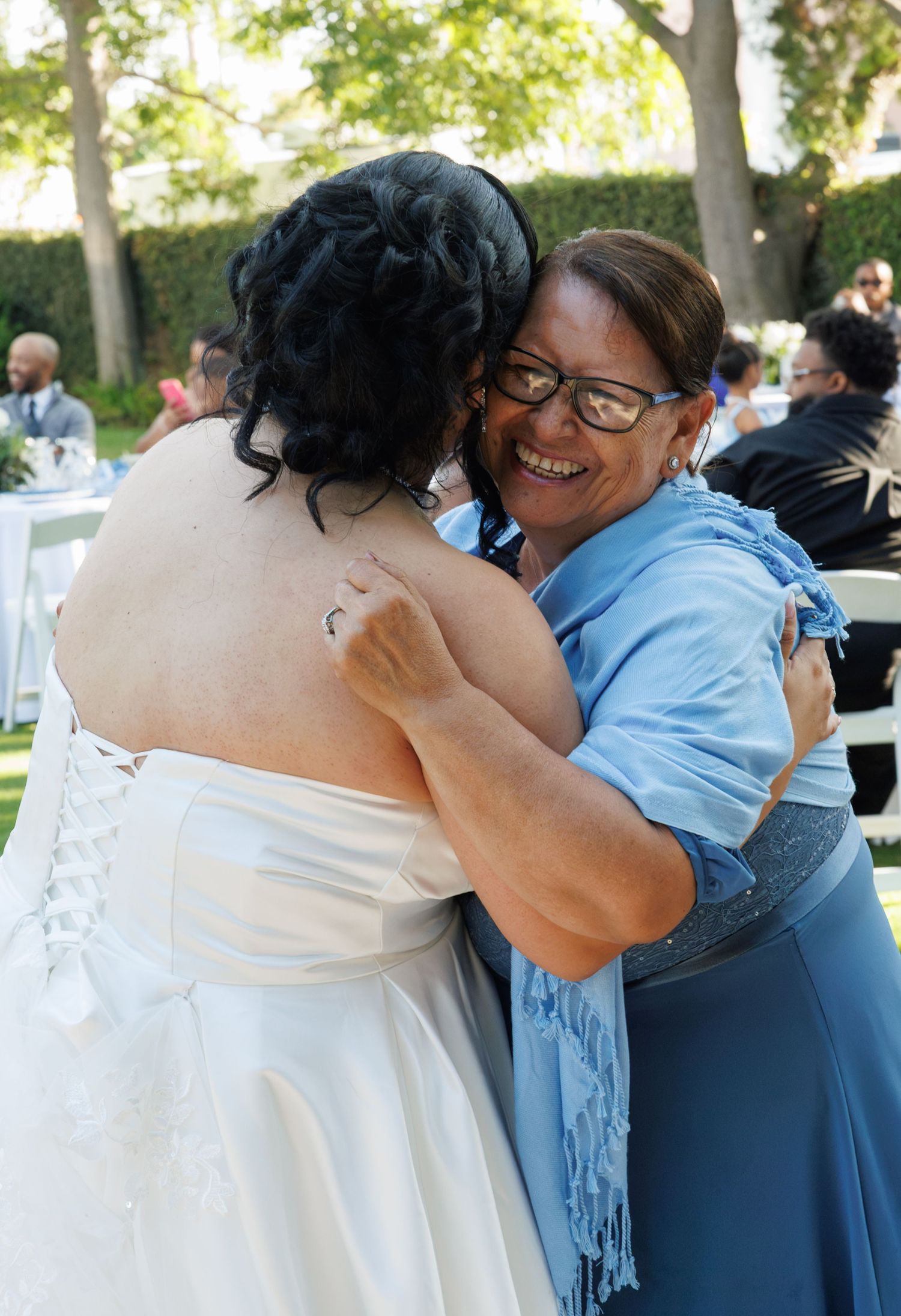 Smiling woman in blue shawl embraces bride in white lace gown at outdoor wedding.