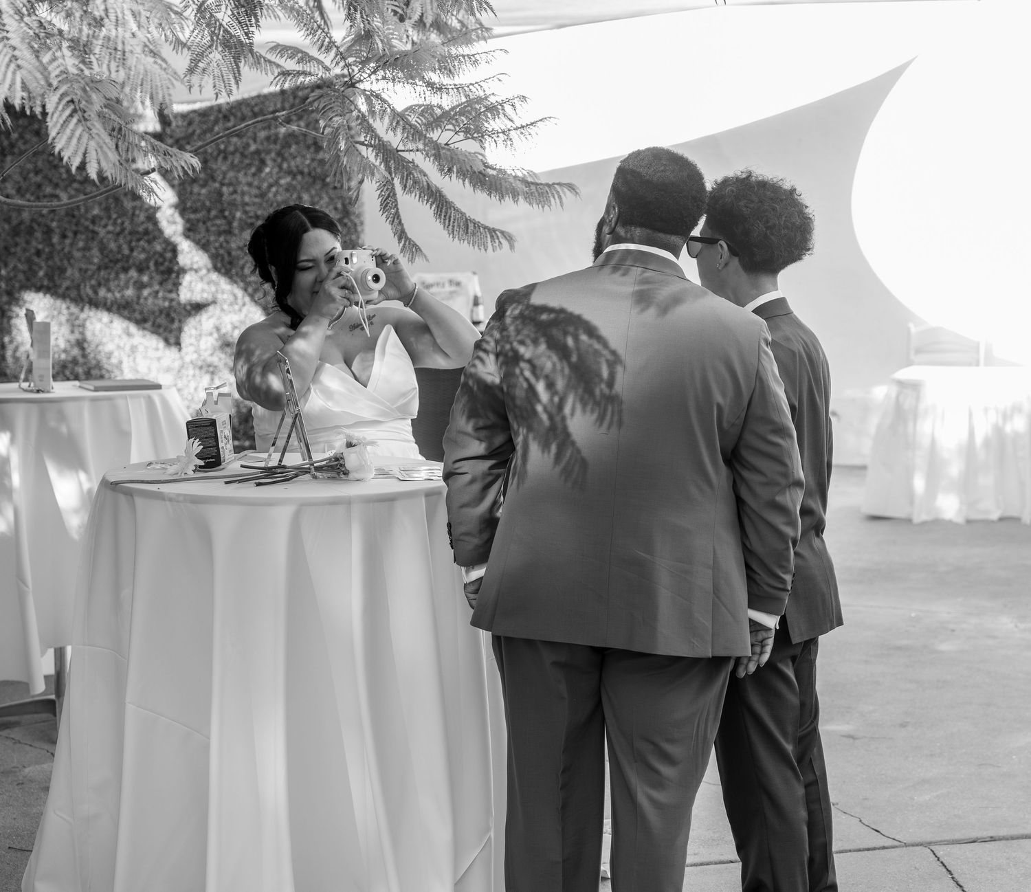 Bride takes a selfie with guests at an outdoor wedding reception with white tablecloths.