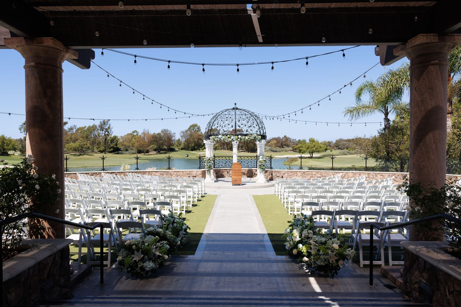 Elegant outdoor wedding ceremony setup with white chairs, string lights, and a gazebo overlooking a scenic golf course.